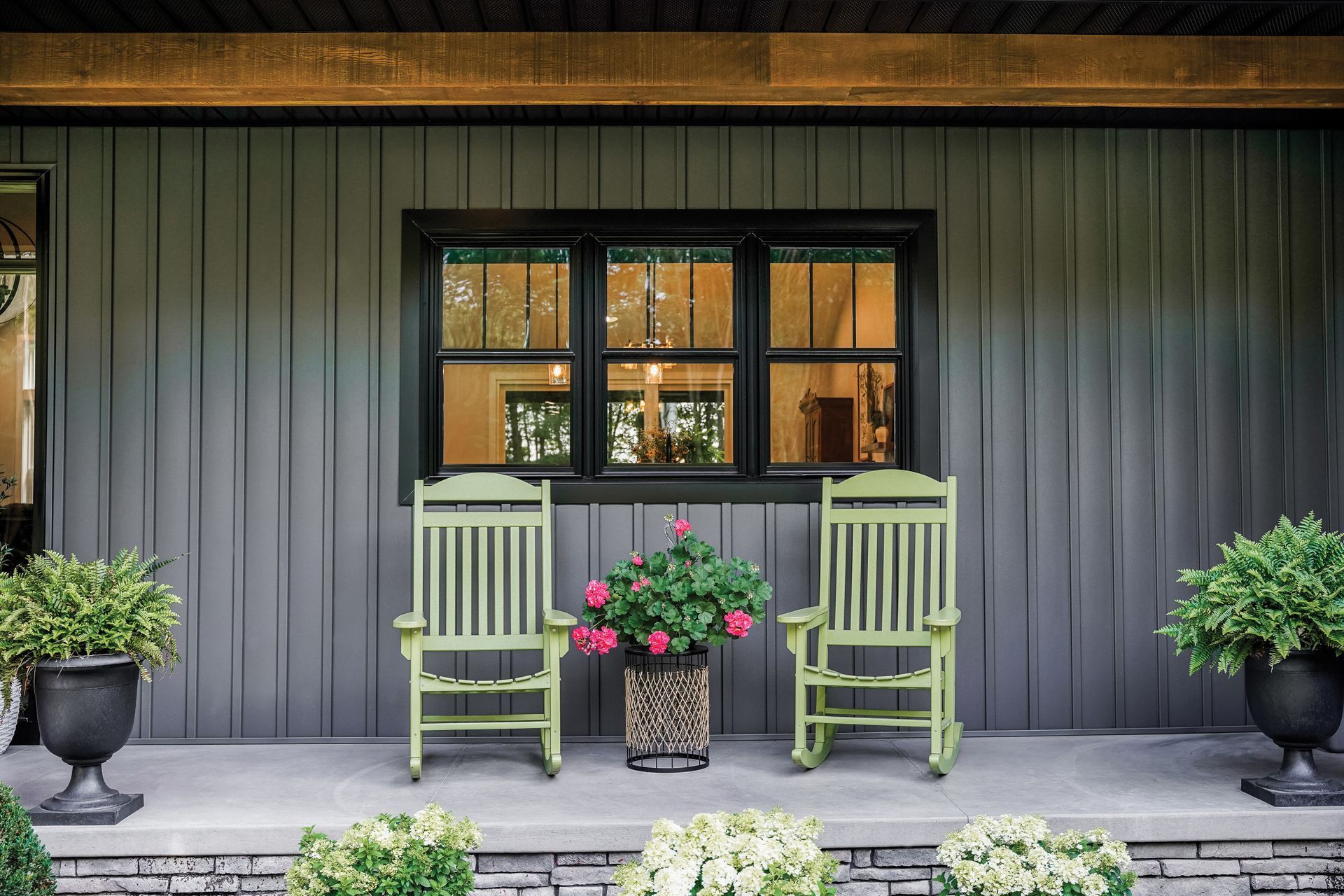 Two light green rocking chairs on a gray porch with vertical siding, a central window, and potted plants.