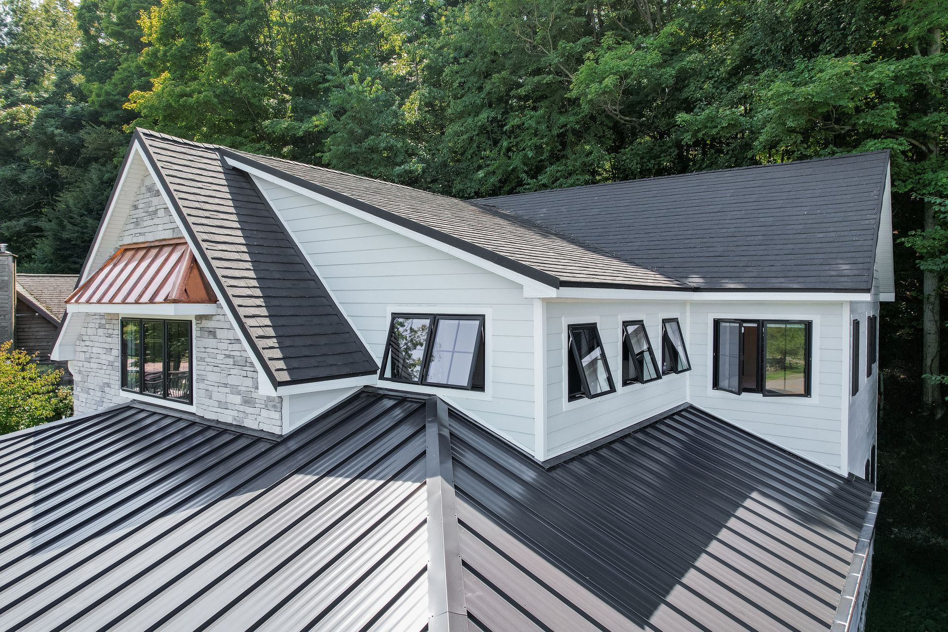 Aerial view of a house featuring a mix of shingle and metal roofing, white horizontal siding, and dark-framed windows.