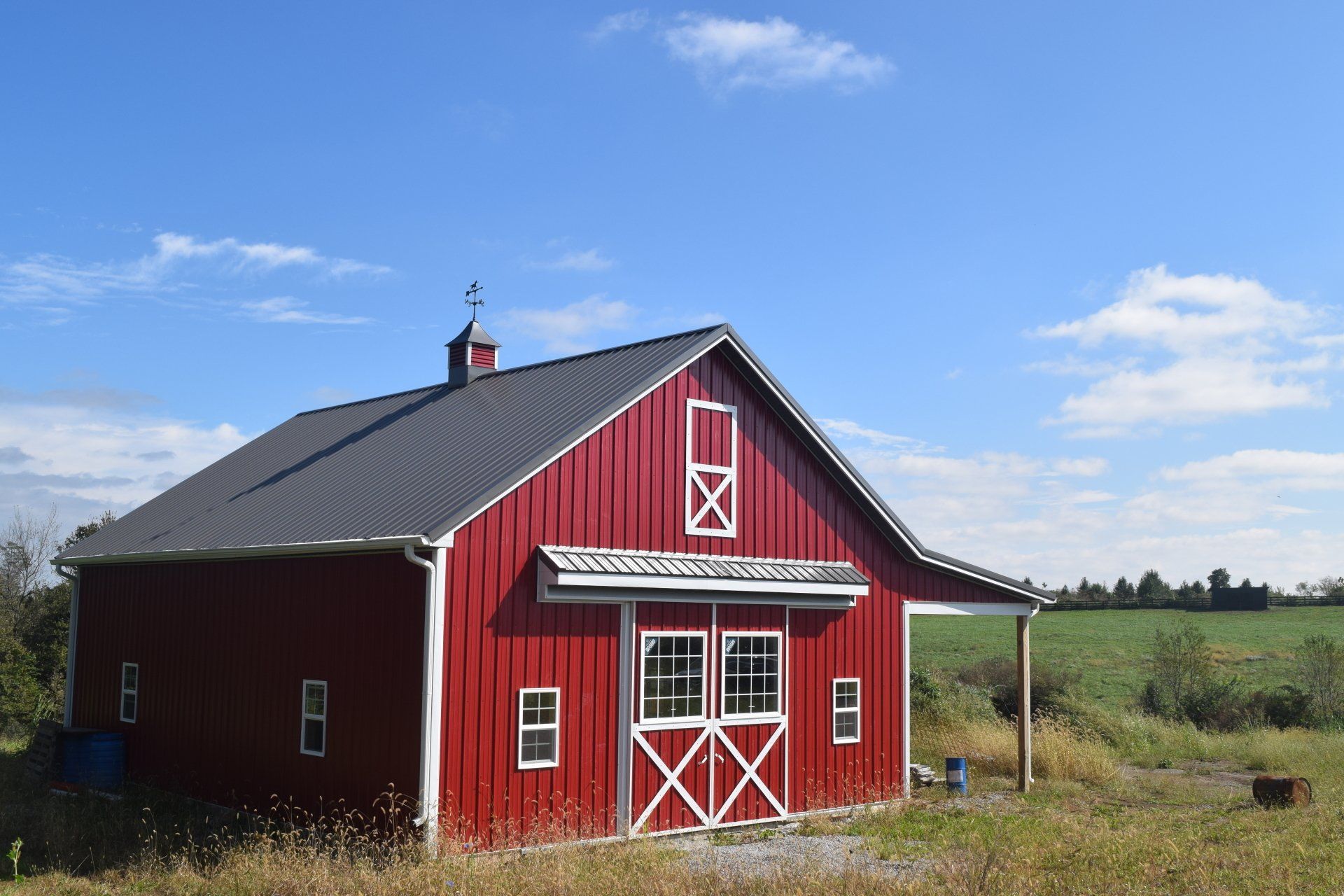 A small red barn with a gray metal roof, white trim, and double doors, situated in a field under a clear blue sky.