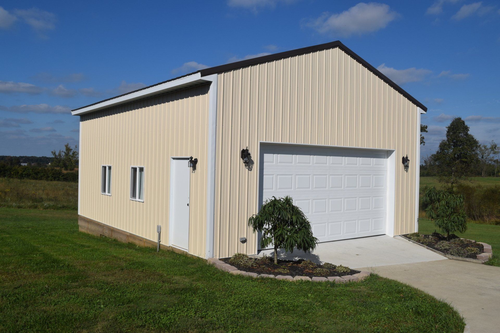 Beige metal garage with a white roll-up door, side entry, and two windows, set in a grassy field under a blue sky.