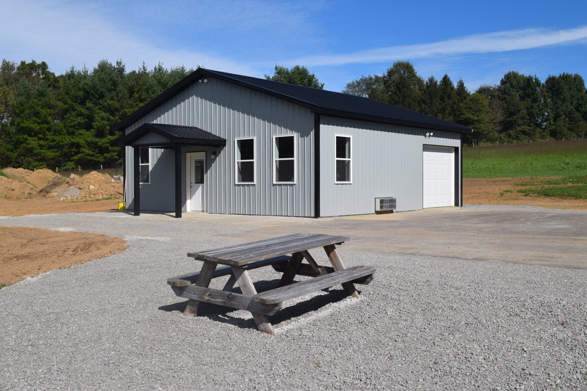 A grey metal building with a black roof and white door, situated on a gravel lot with a wooden picnic table in front.