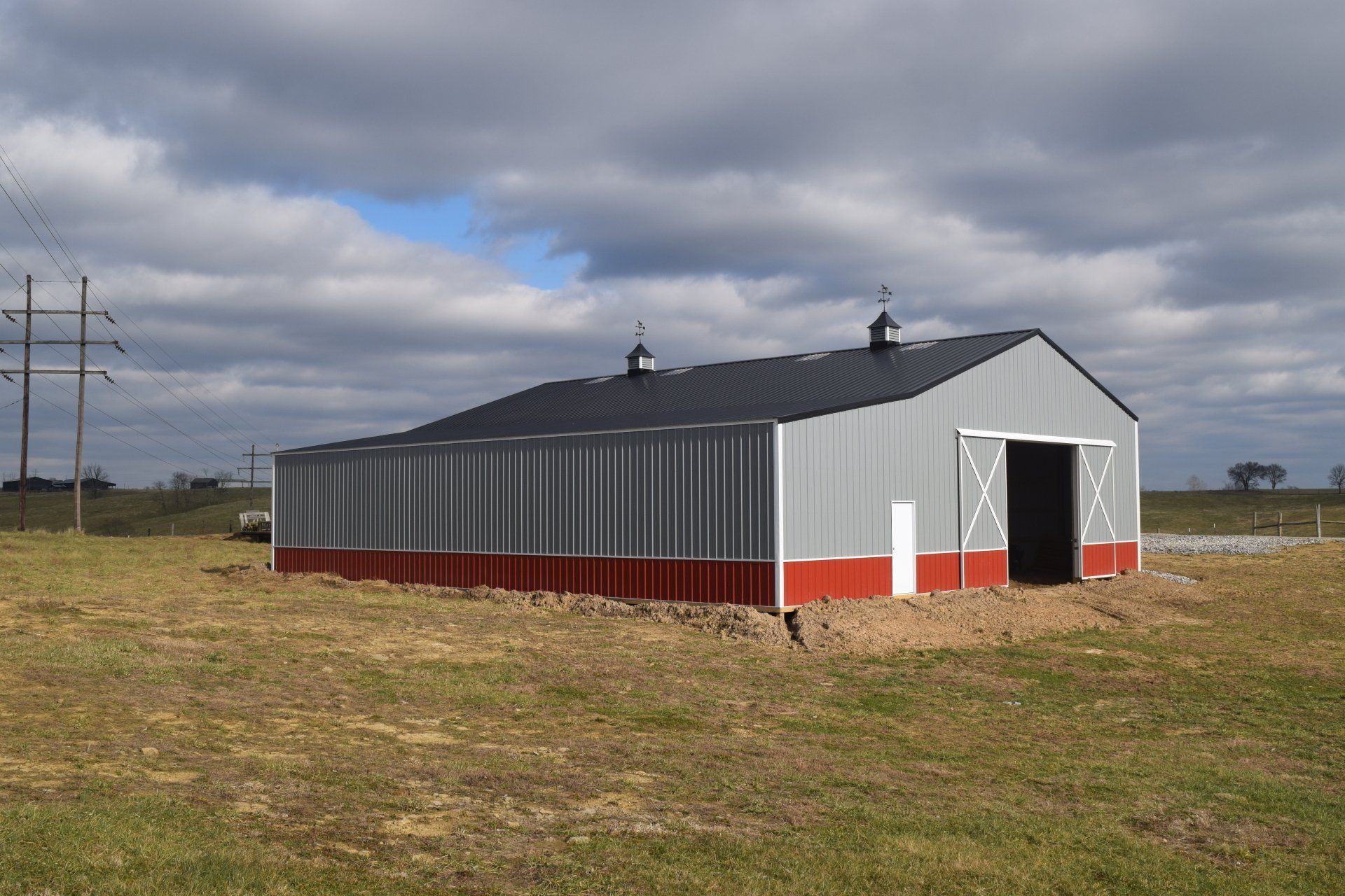 A metal pole barn with gray walls and a red base sits in a grassy field under a cloudy sky.