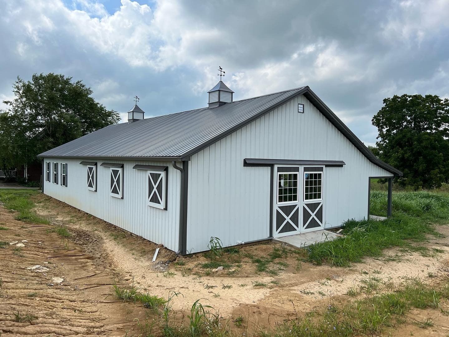A white barn with a grey metal roof and cupolas, set against a cloudy sky with dirt ground in the foreground.