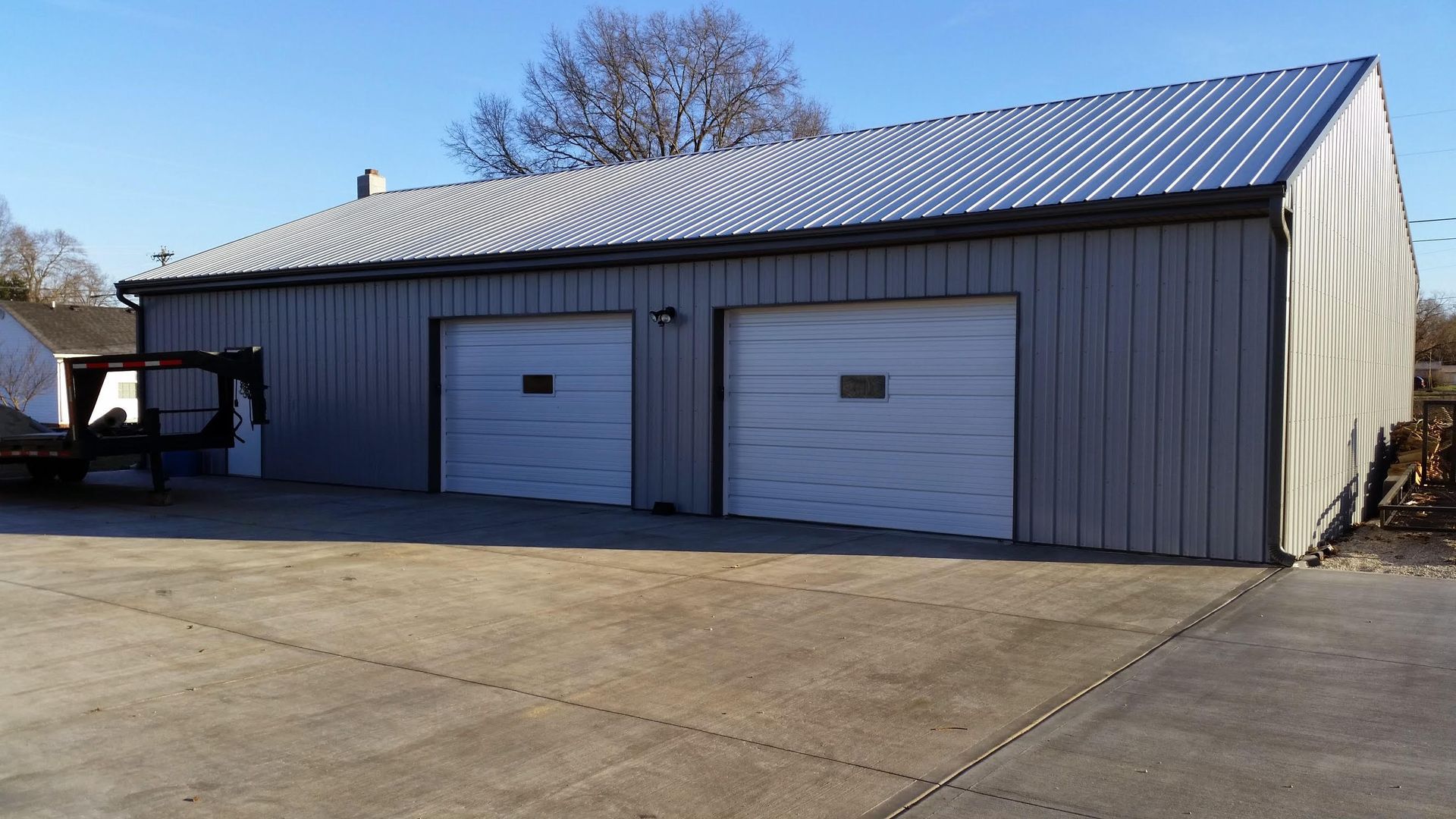 A grey metal warehouse building with two white garage doors, situated on a large concrete driveway under a clear sky.