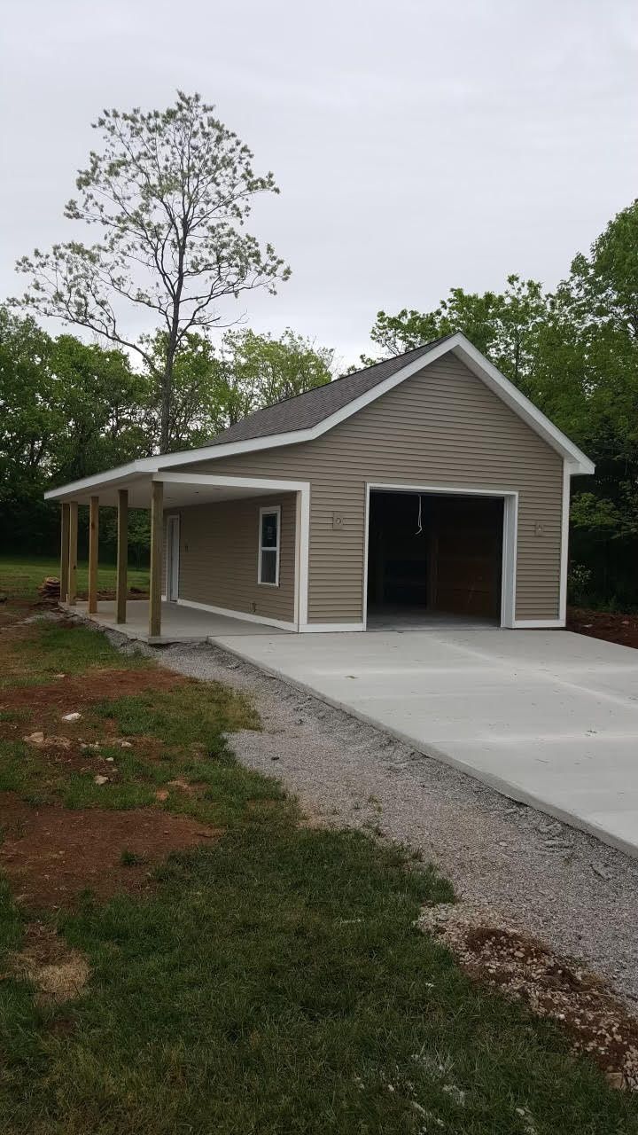 A tan garage with white trim and an attached side porch, sitting on a concrete pad surrounded by trees and grass.