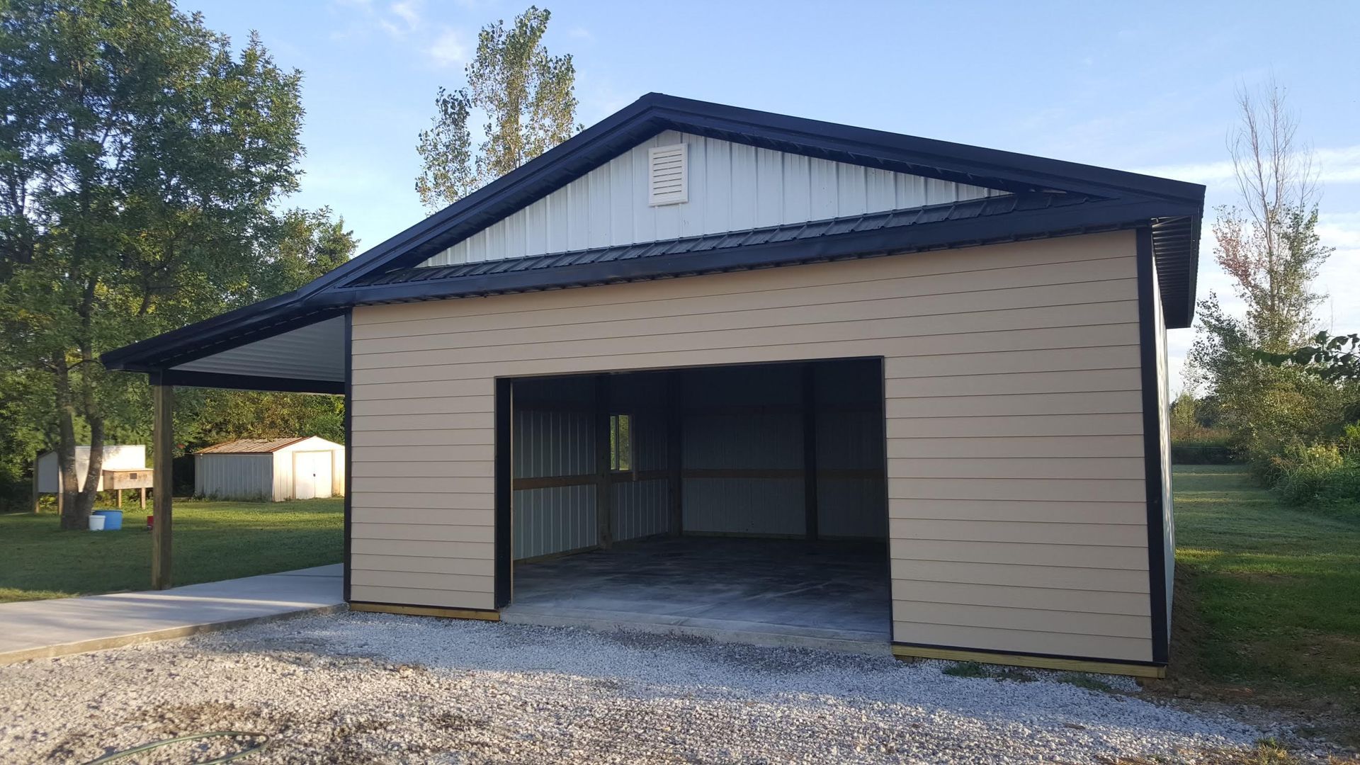 A tan metal garage with a black roof and an open entrance, featuring a side lean-to shelter on a gravel lot.