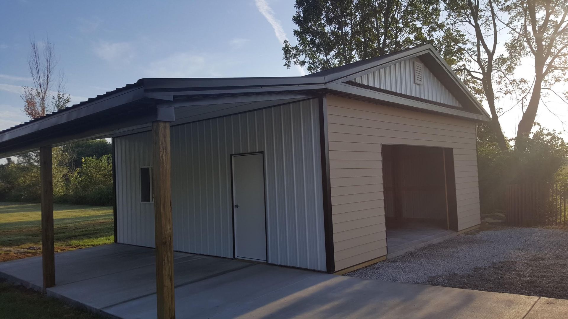 A beige and white metal garage with a covered side carport on a gravel and concrete pad in a grassy, sunlit yard.