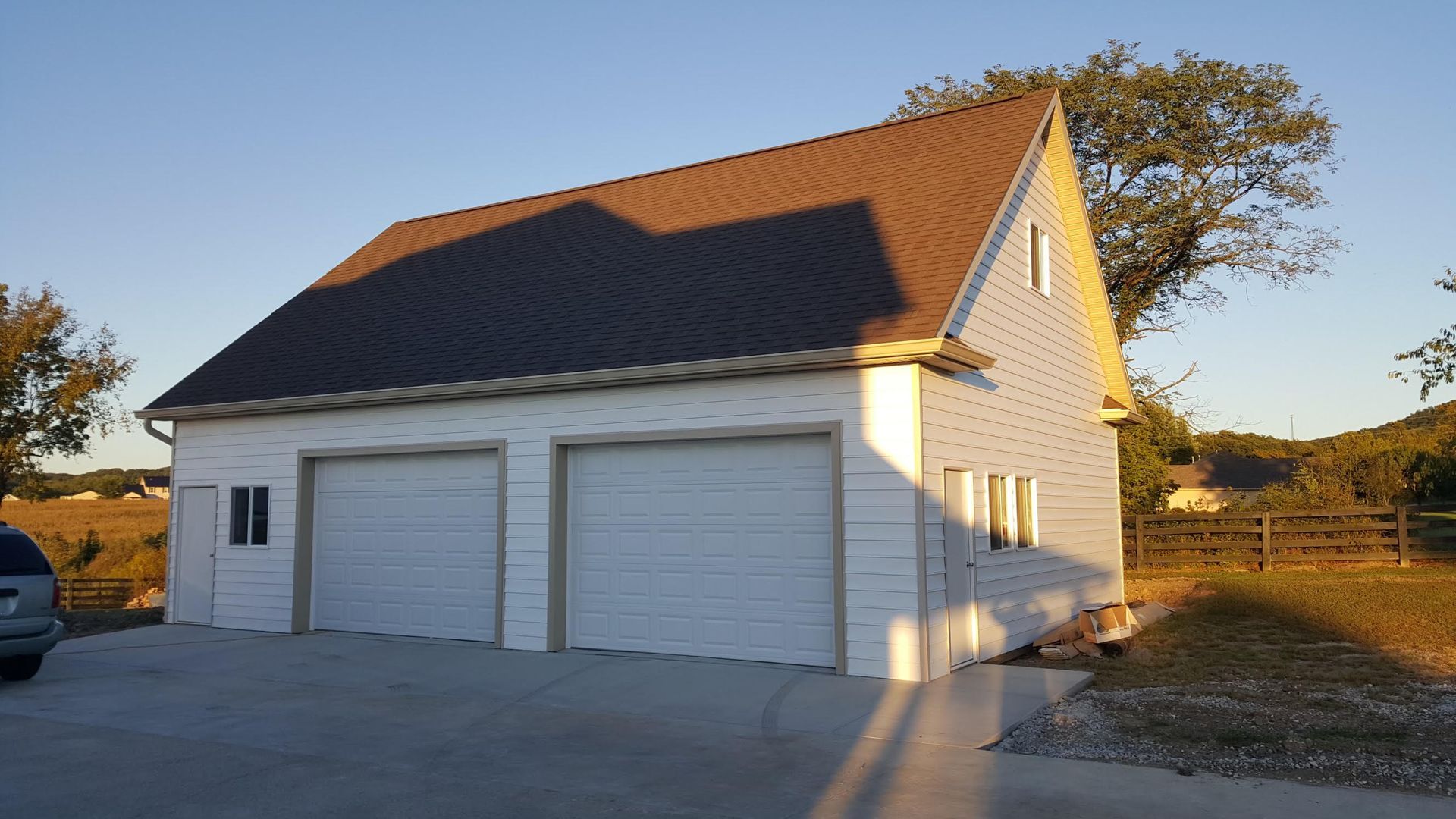 A white detached two-car garage with a brown metal roof sits on a concrete pad in a rural, sunny setting.