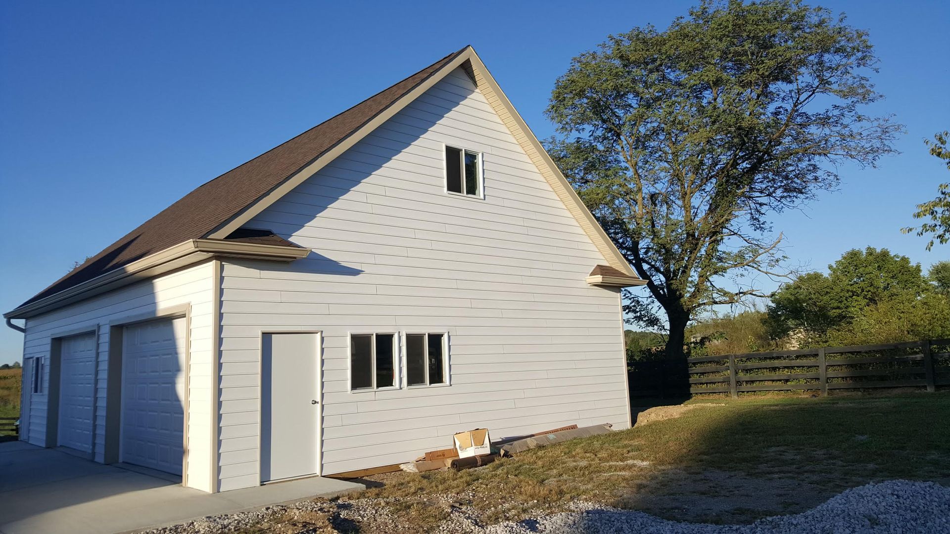 A white two-story detached garage with two bays and a side door, situated on a grassy lot next to a large tree.