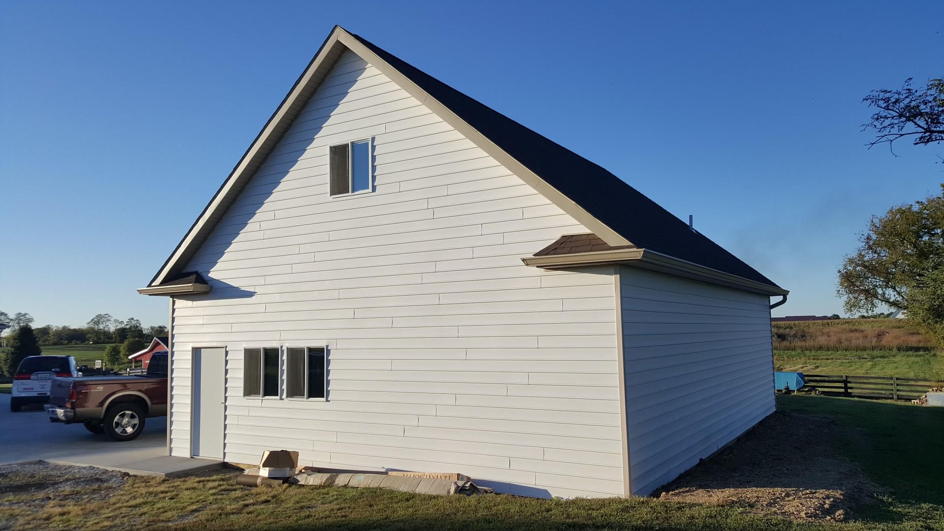 A white, two-story vinyl-sided building with a dark roof and a small window in the attic, set in a rural, sunny yard.