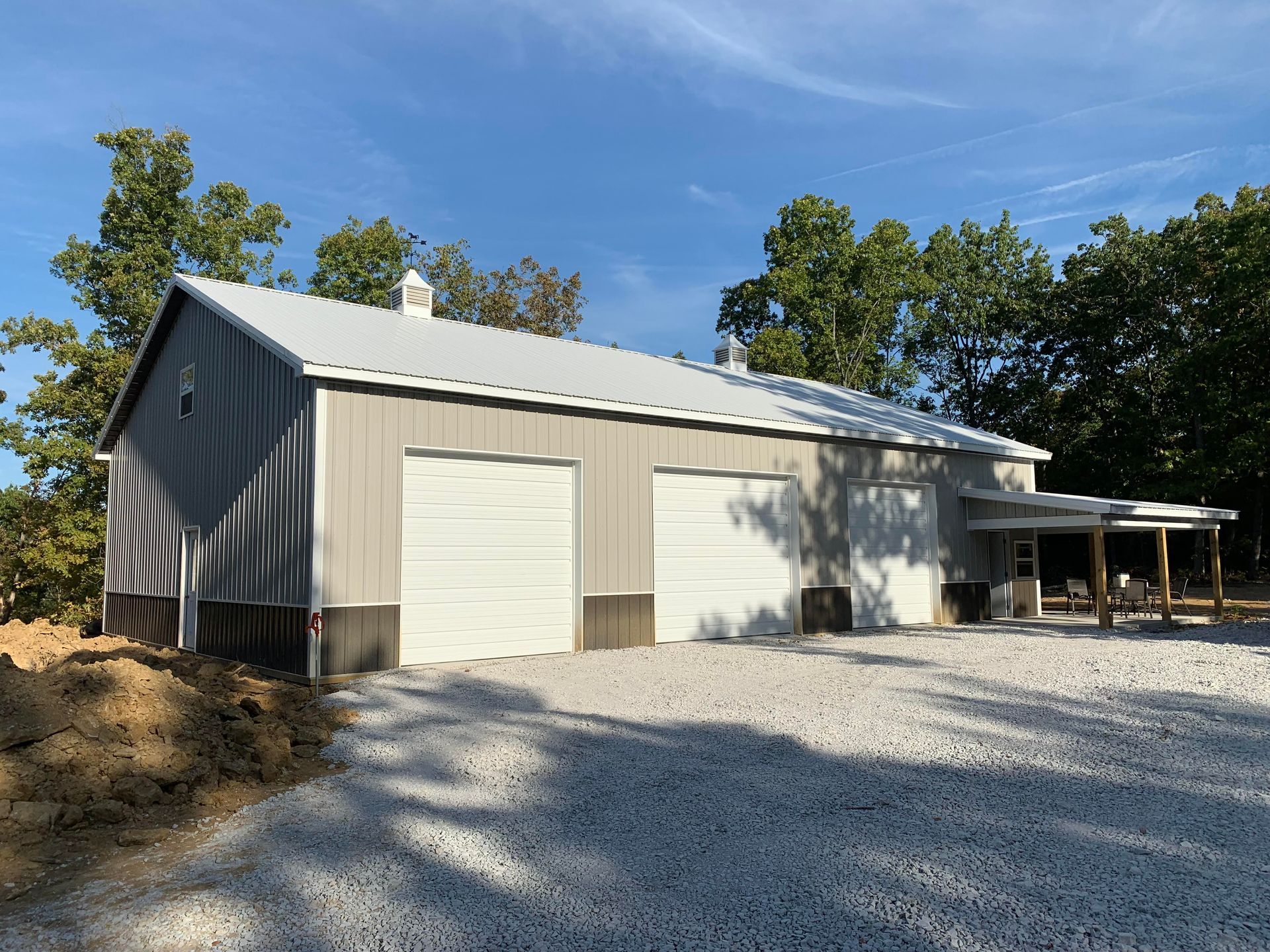 A light gray metal workshop building with three white garage doors, set on a gravel lot next to trees under a blue sky.