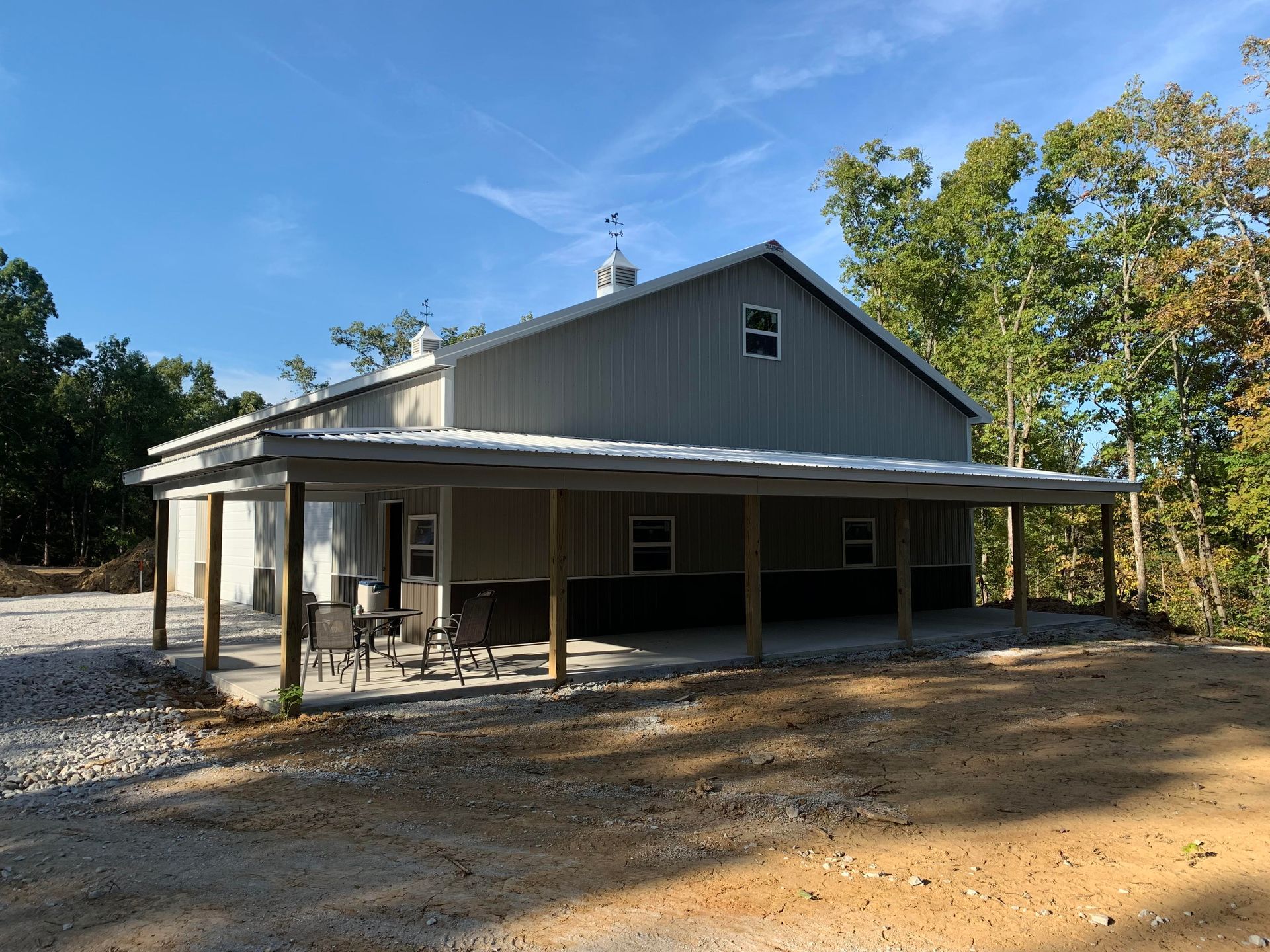 A gray metal pole barn with a full-length covered porch, surrounded by trees on a partially gravel, partially dirt lot.