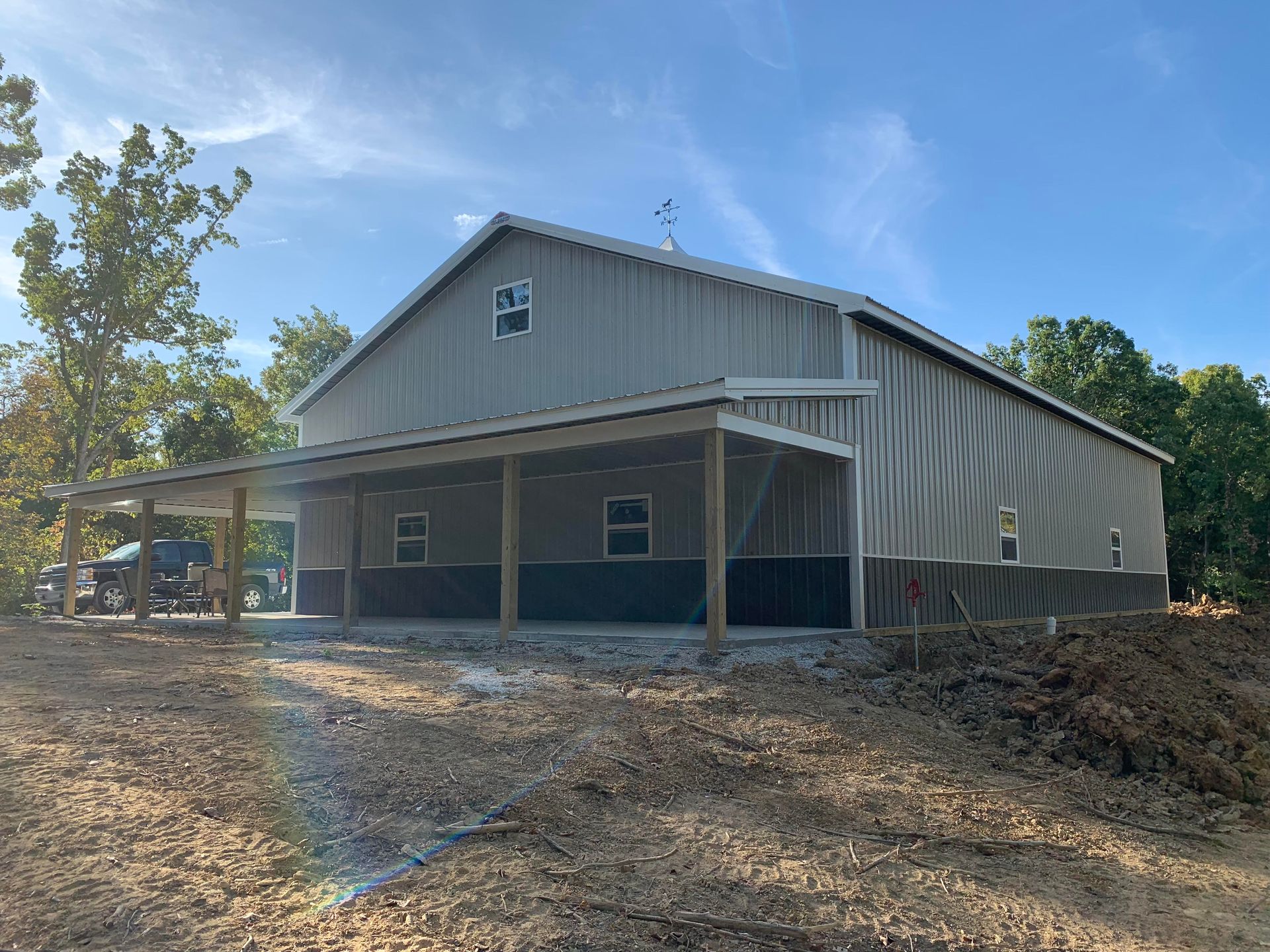 A large, metal-sided barn-style building with a covered porch, set on a gravel lot under a bright blue sky.