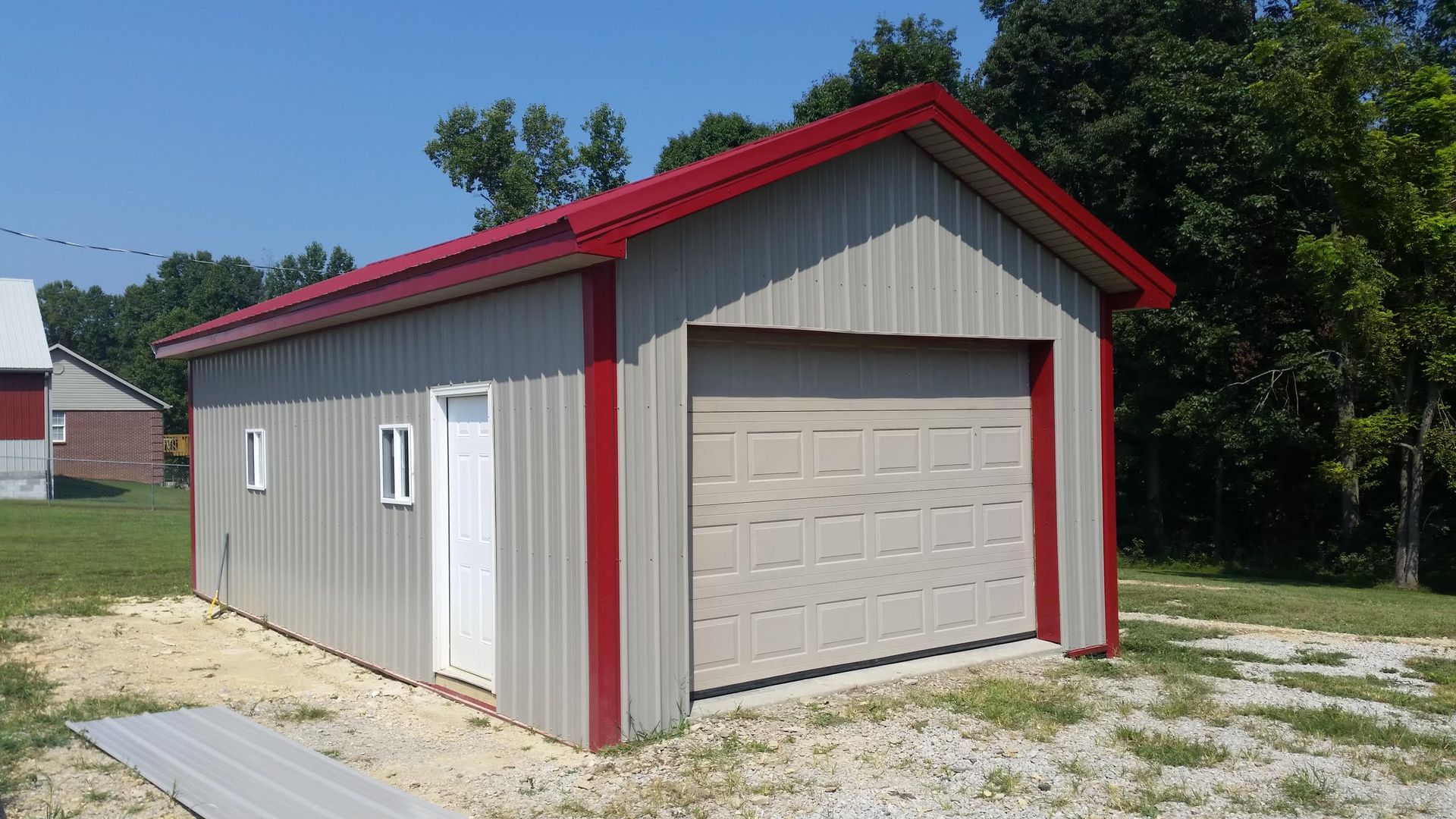 A tan metal garage with red trim, a single white entry door, and a white overhead garage door on a gravel lot.