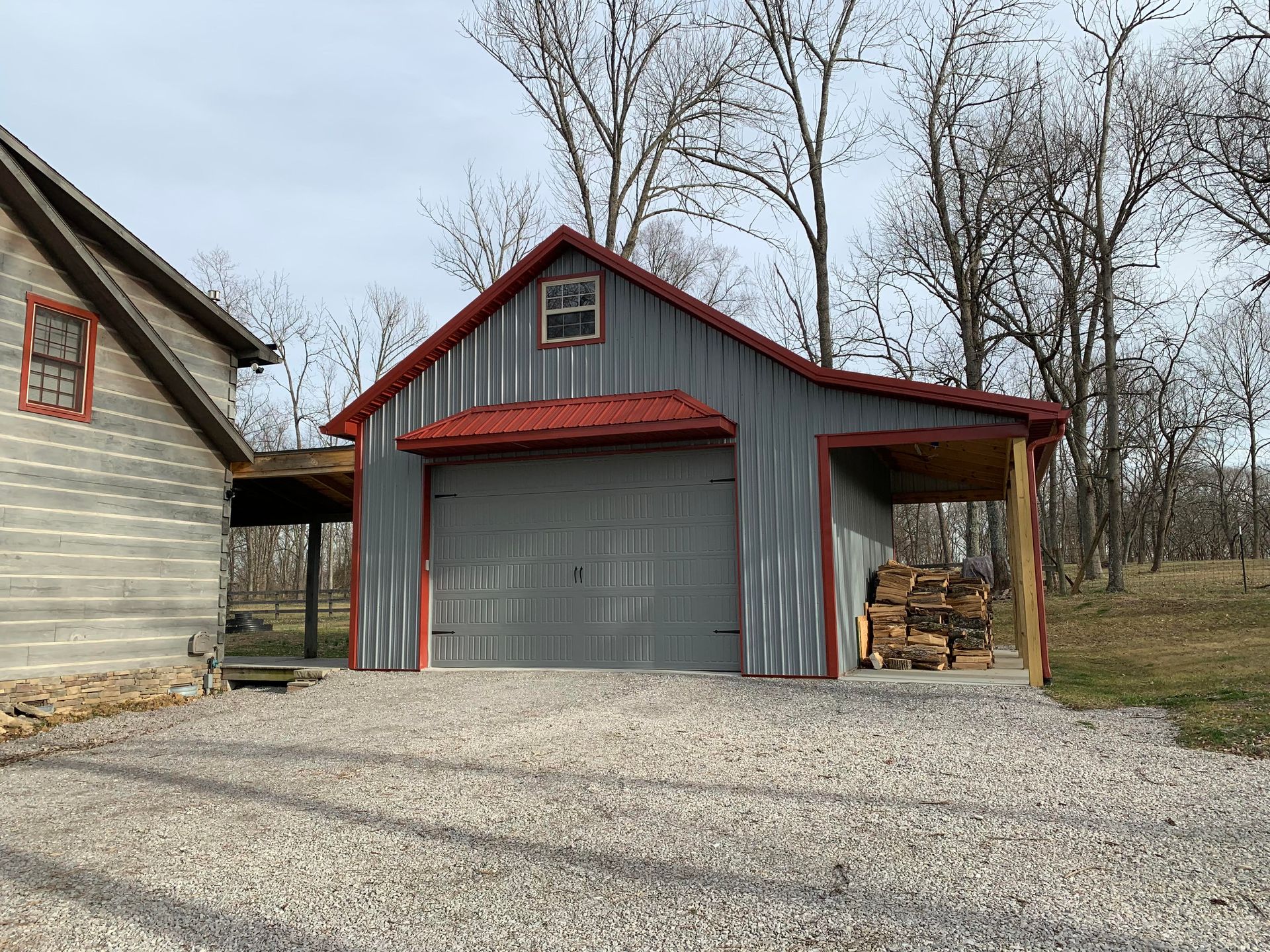 A grey metal garage with a red roof, a matching lean-to with stacked firewood, and a log cabin on a gravel driveway.