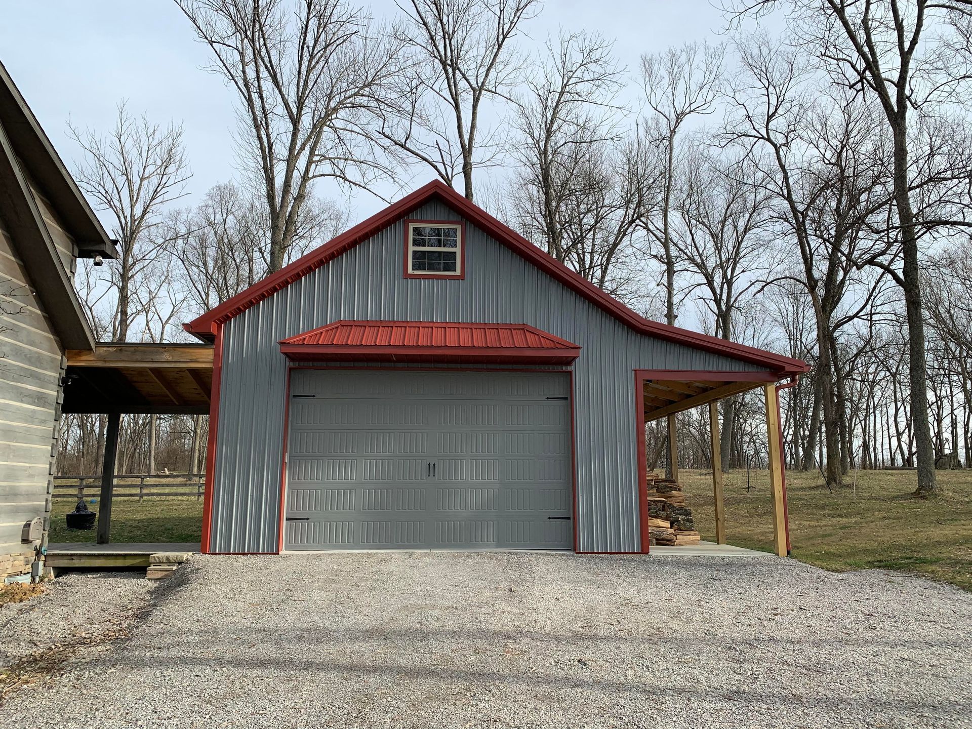 A grey metal garage with a red roof, featuring a central garage door, a small upper window, and side lean-to structures.