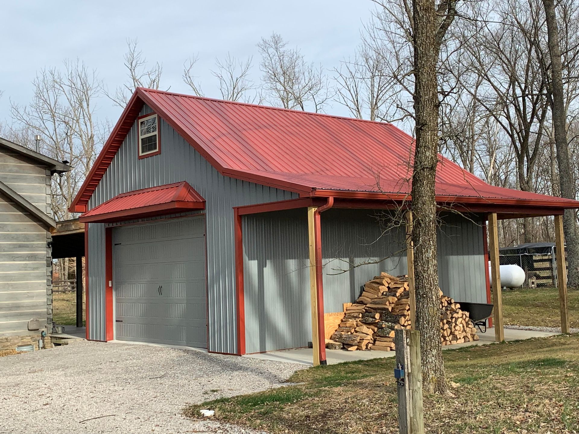 A grey metal garage with a red roof, featuring a side shed area filled with a large stack of firewood in a wooded yard.