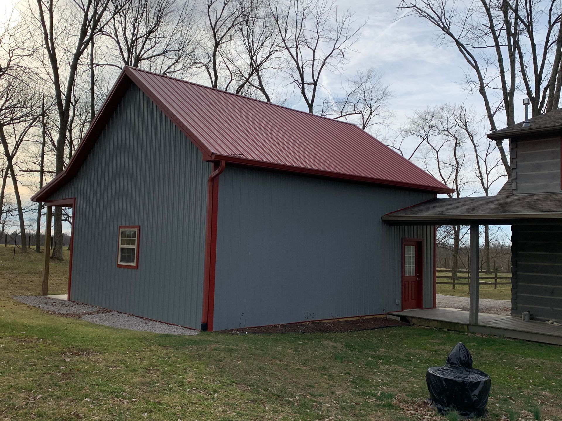 A grey metal barn with a red roof and red trim stands in a grassy clearing with trees in the background.