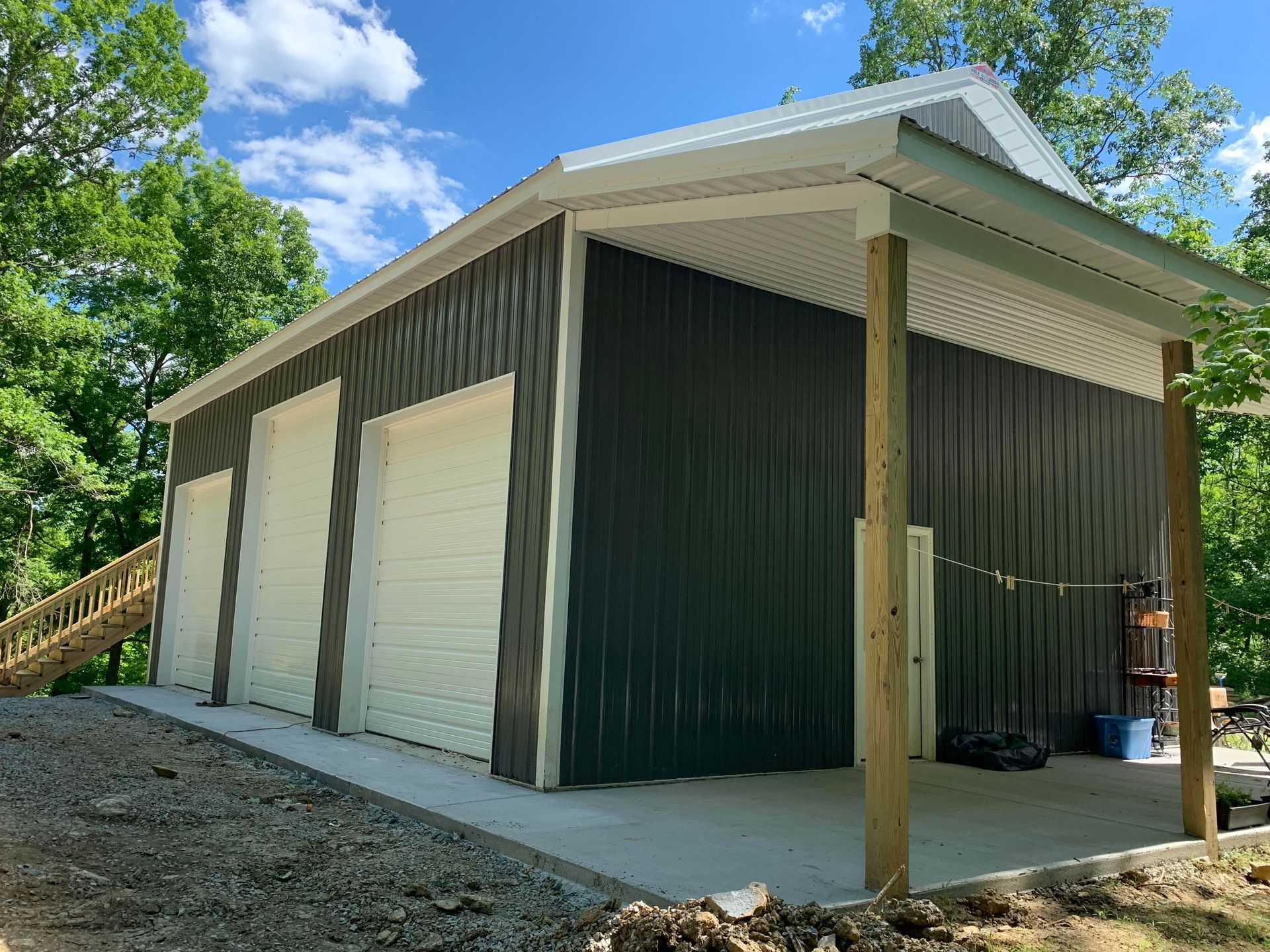 A charcoal-colored metal garage with two white bay doors, a concrete porch, and wooden support posts in a wooded setting.