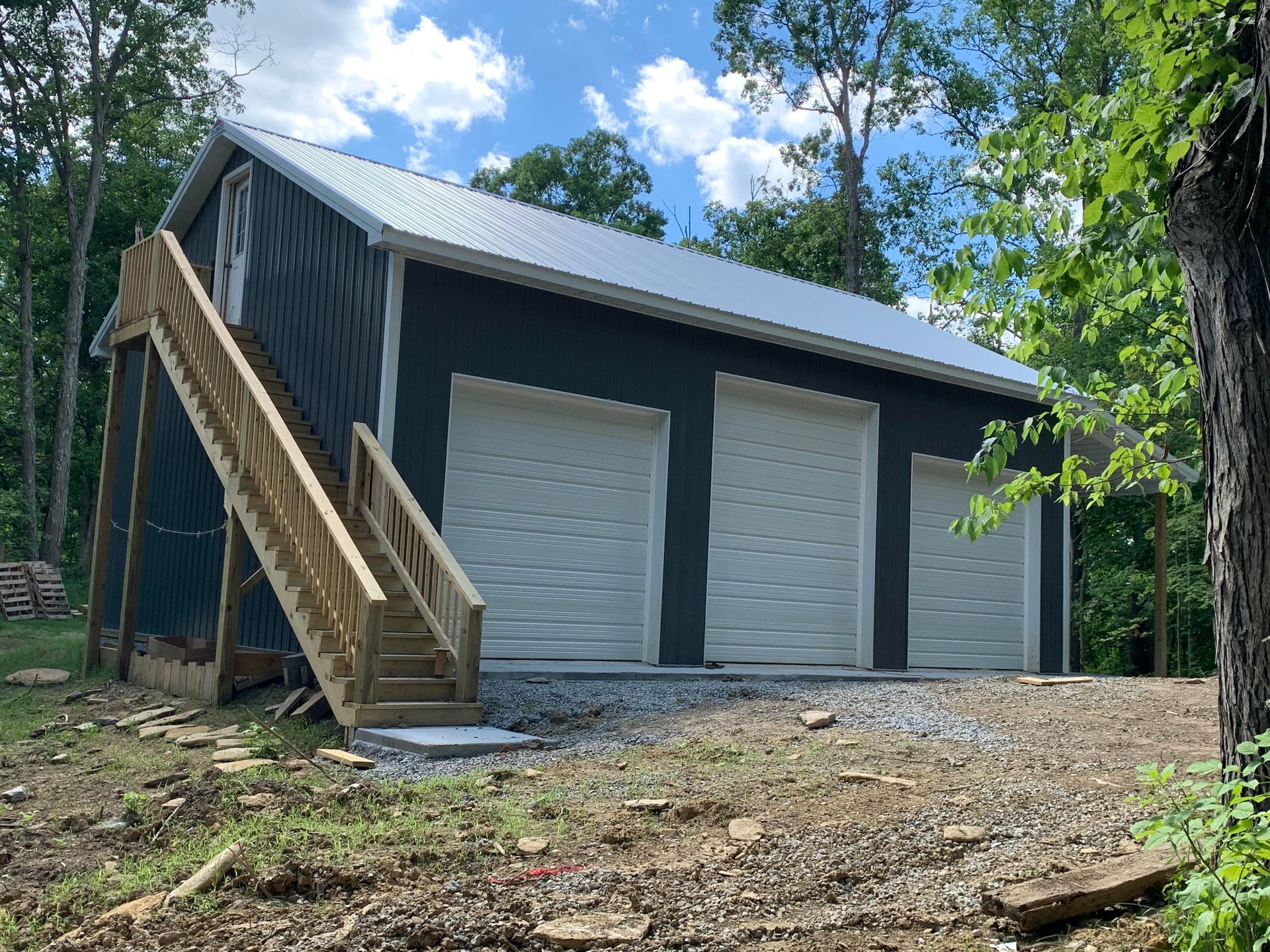 A detached, dark blue garage with a metal roof and three white doors, featuring an exterior wooden staircase to an attic.