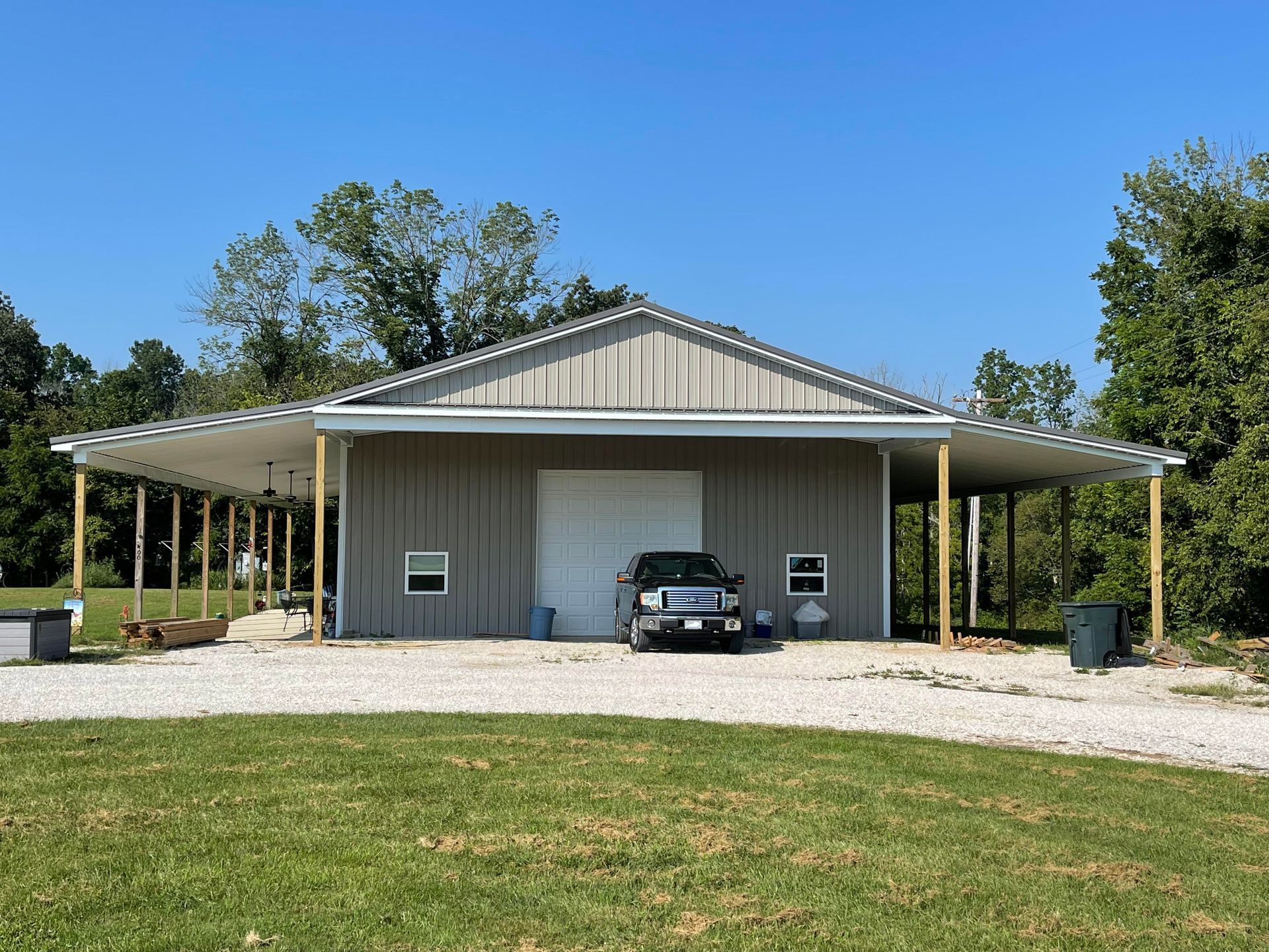 A grey metal barn with a covered porch on both sides, a white garage door, and a black truck parked in front.