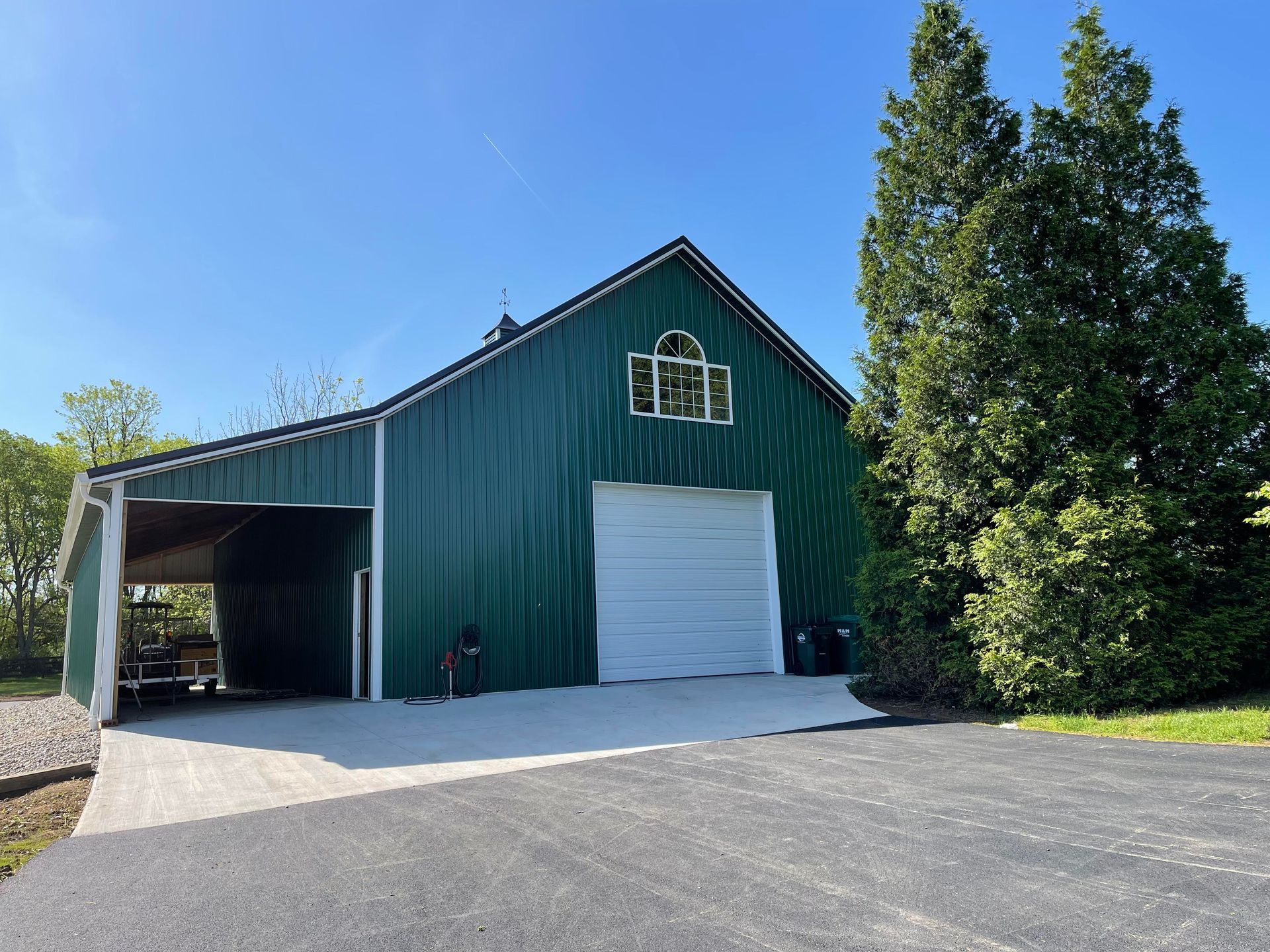 A green metal barn with a white garage door, an attached side carport, and two tall evergreen trees against a blue sky.