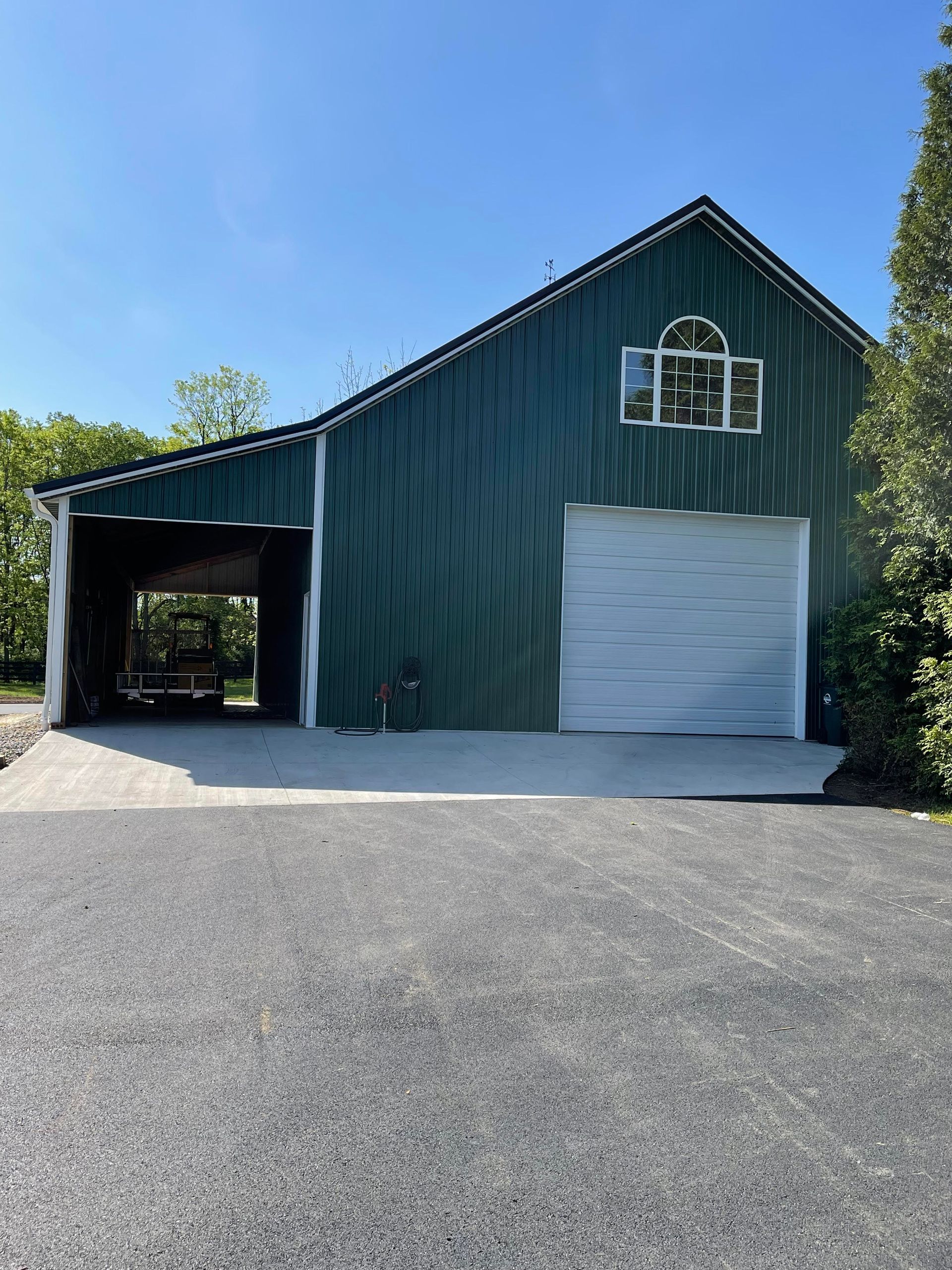A green metal barn featuring a white roll-up door, a side open-air bay, and a decorative arched window on a gravel lot.