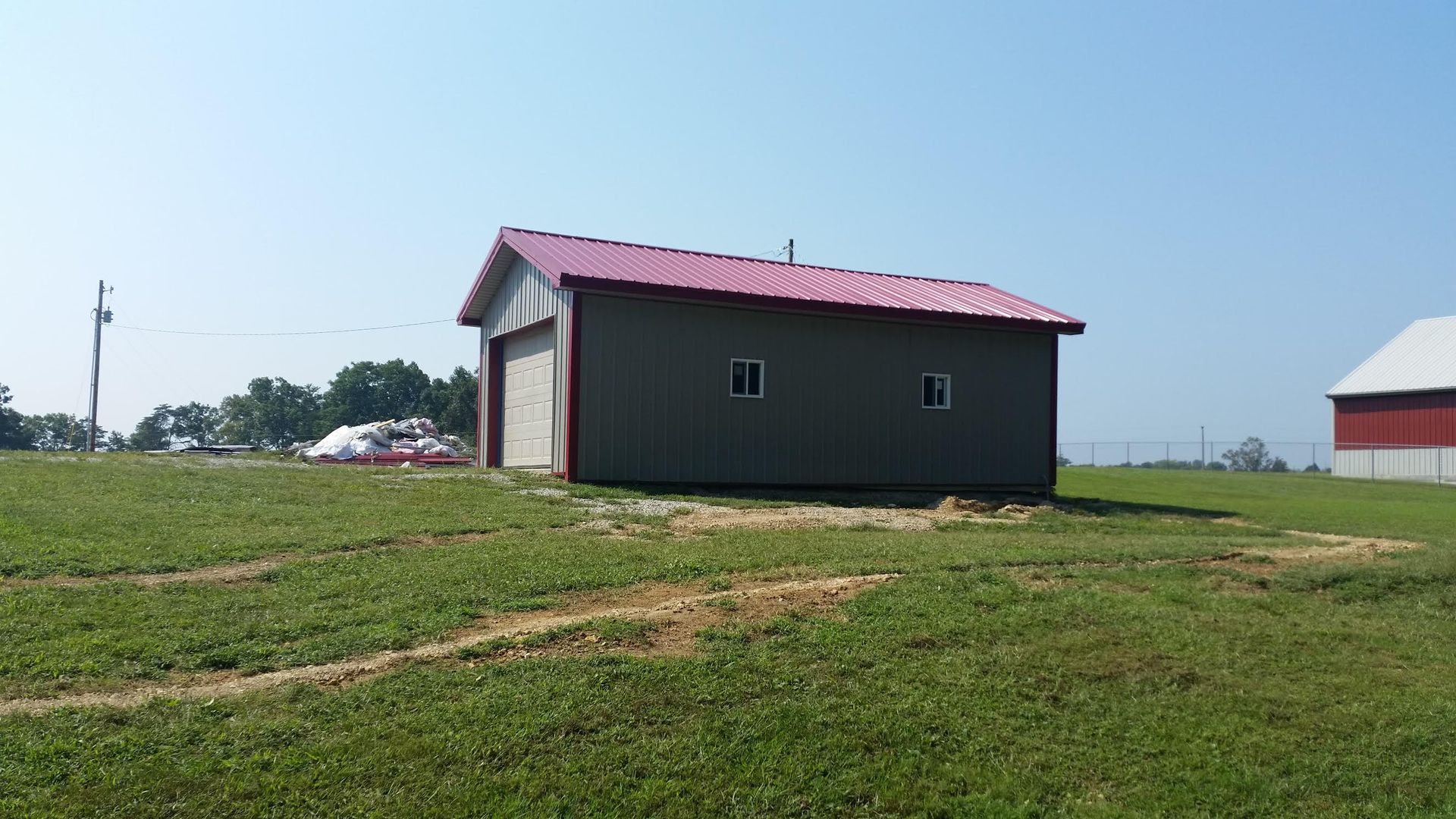 A grey pole barn with a red metal roof sits on a grassy field under a clear blue sky, with a red barn in the distance.