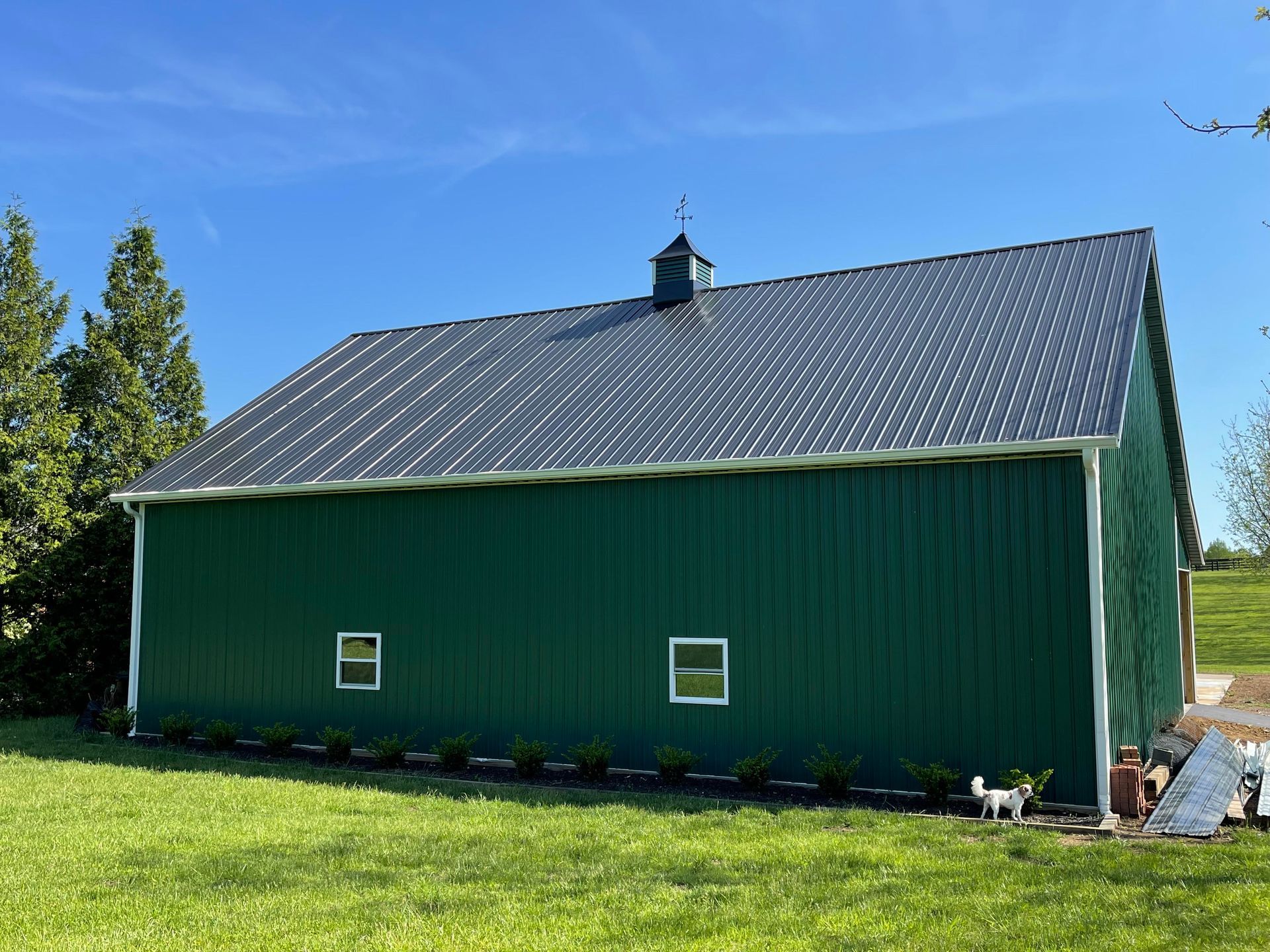 A dark green metal-sided barn with a dark gray gabled roof and a small cupola, set in a grassy field under a blue sky.