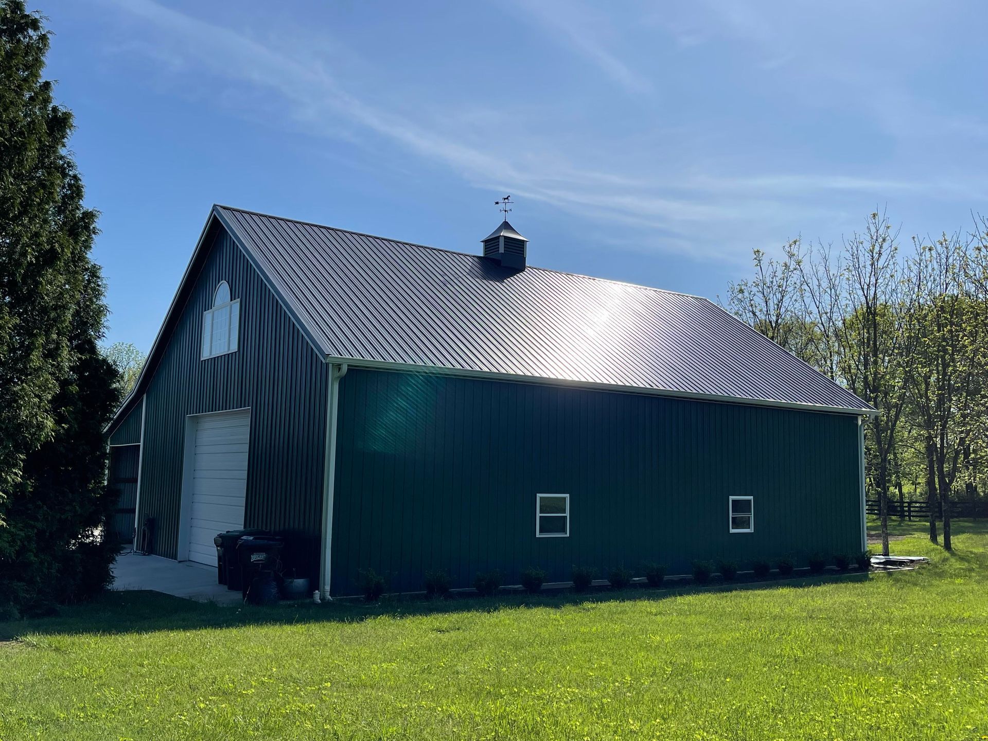 A dark green pole barn with a metal roof and a small cupola sits on a green lawn under a clear blue sky.