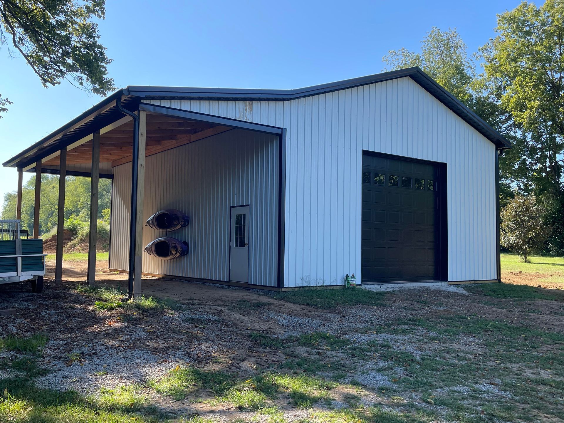 A white metal pole barn with a black garage door and an open-sided lean-to attached on the left, set on a gravel lot.