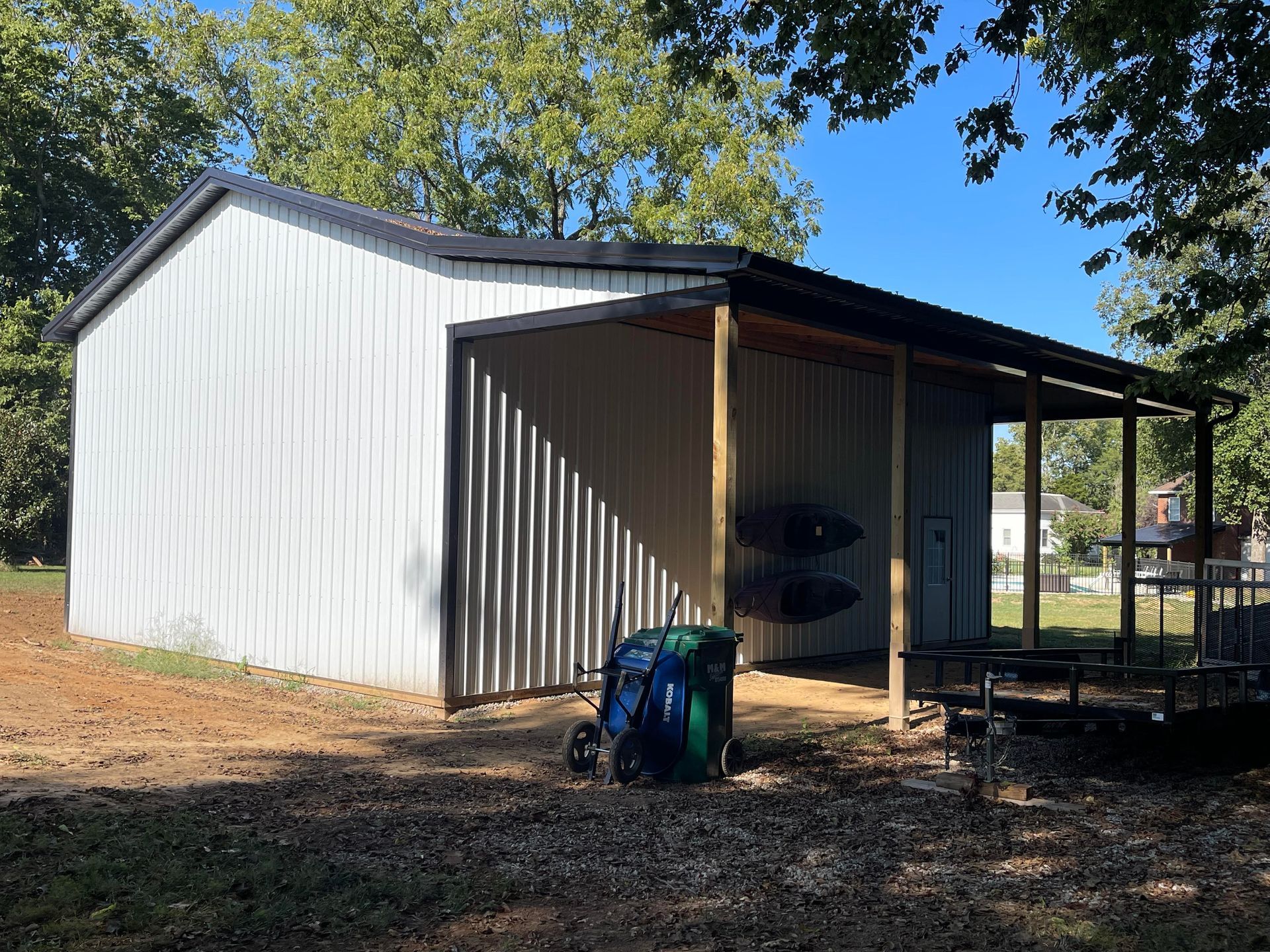 A white metal shed with a side carport containing kayaks, standing on a dirt lot surrounded by trees.