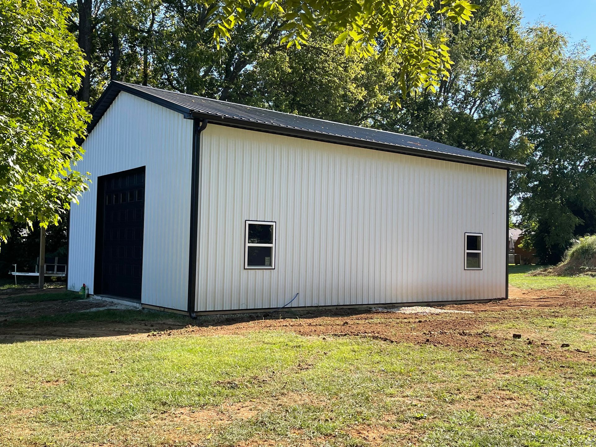 A white metal workshop with black trim and two windows sits on a grassy lot with trees in the background.