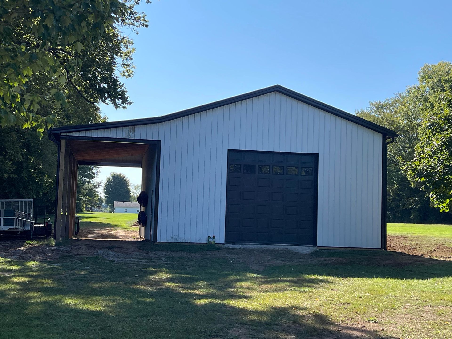 A white, metal-sided garage with a dark door and an open-sided carport, situated on a grassy lot with trees.