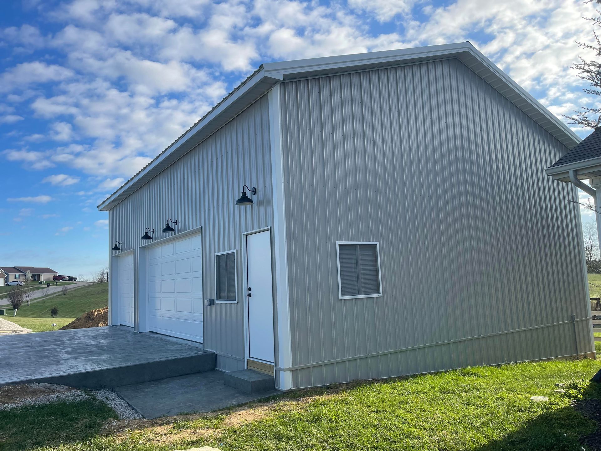 A light gray, metal-sided building with two garage doors and an entry door, set in a grassy yard under a blue, cloudy sky.