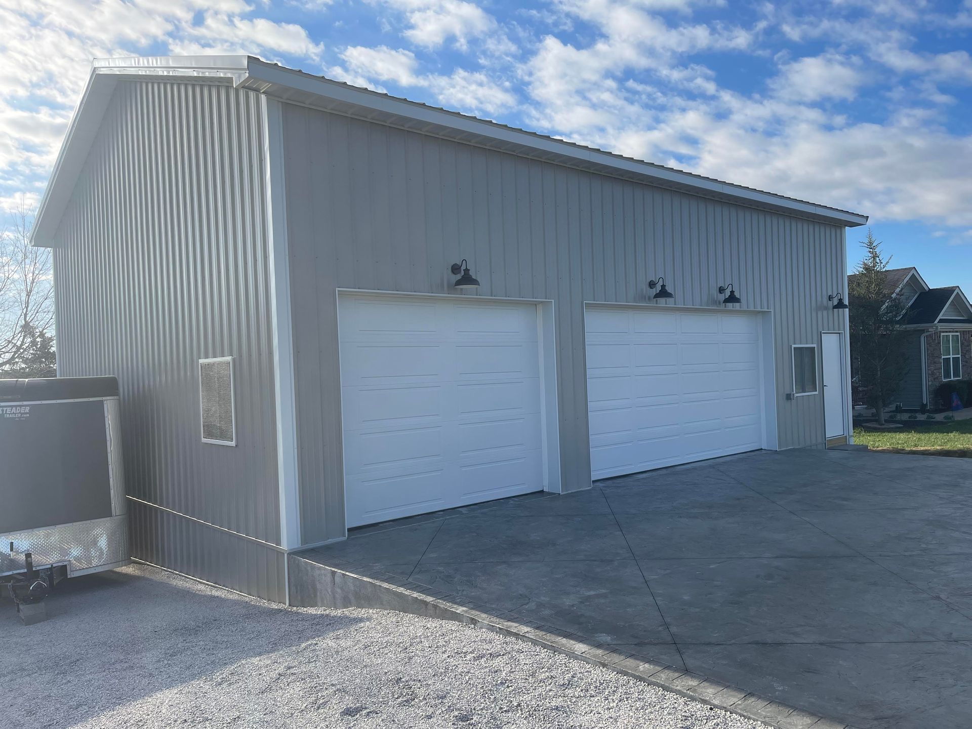 A light gray metal garage building with two white roll-up doors, wall-mounted lamps, and a side door, set on a paved lot.