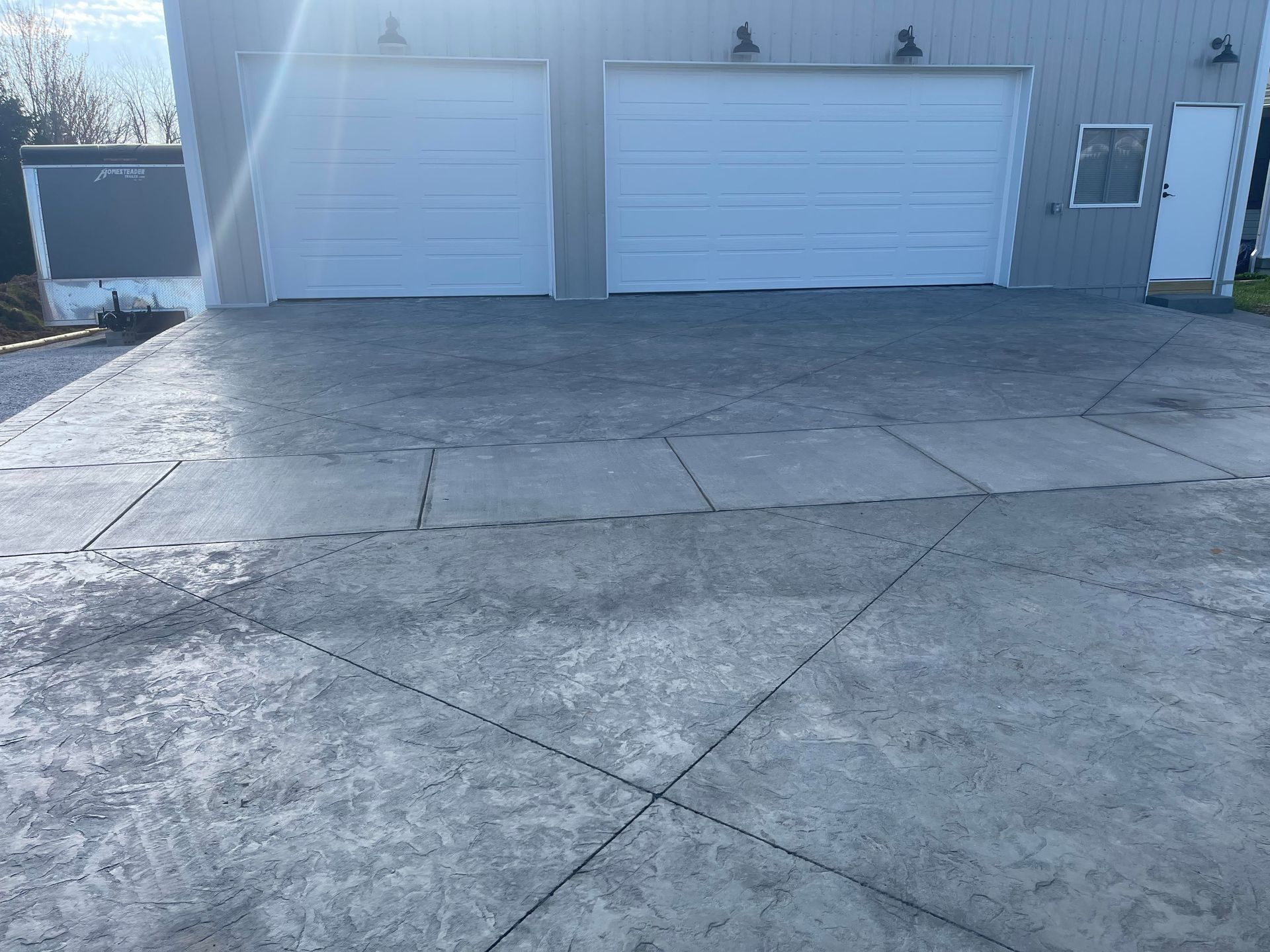 A large, gray stamped concrete driveway leads to a modern metal building with two white garage doors and a side door.