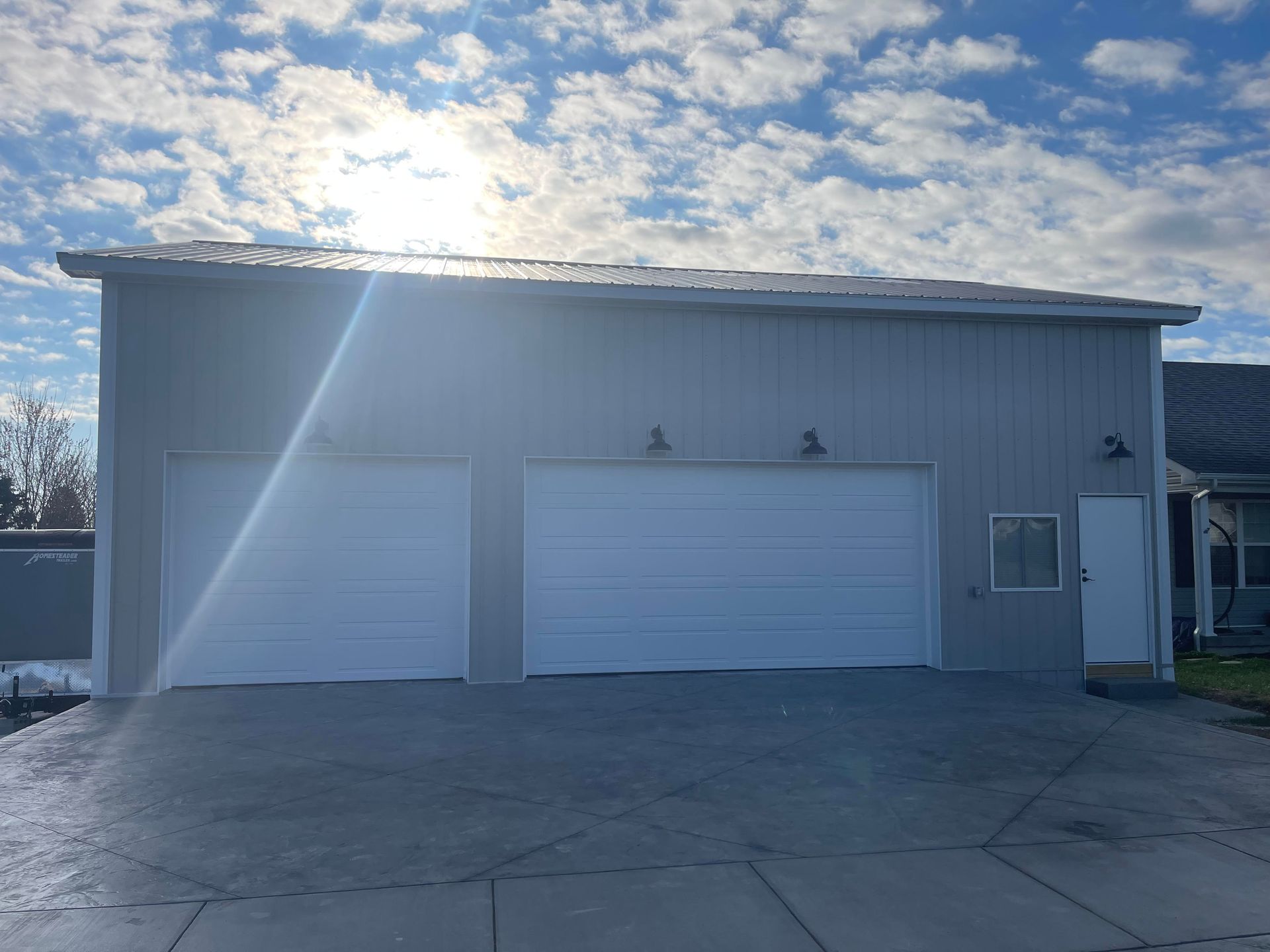 A light grey detached garage with two white doors and a side entrance, set against a blue sky with scattered clouds.