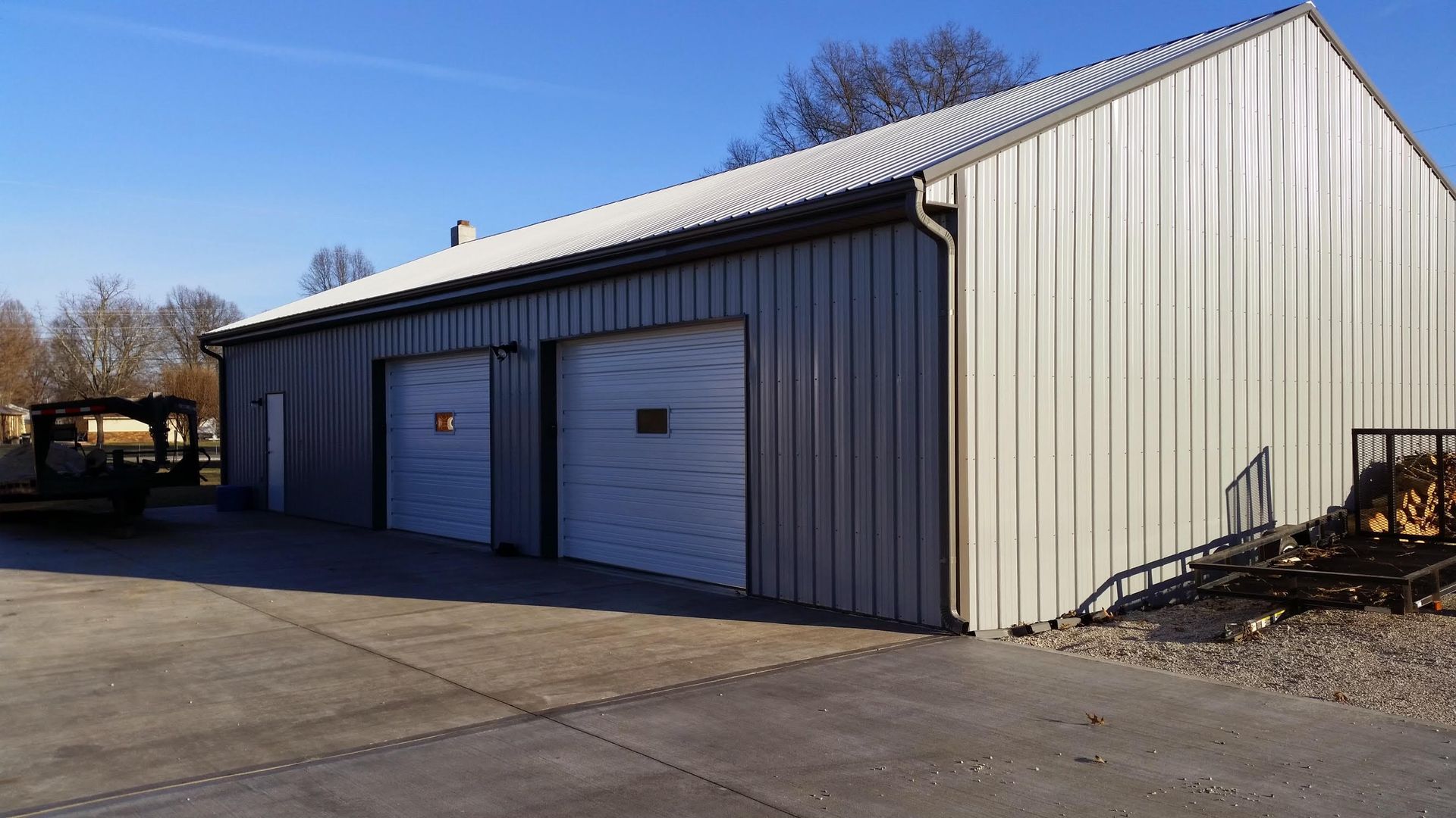 A beige metal workshop building with two white garage doors, situated on a paved lot under a clear blue sky.