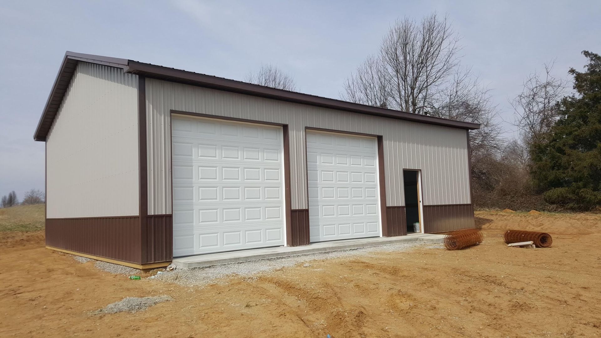 A metal garage with white garage doors and brown trim on a dirt lot under a bright sky.
