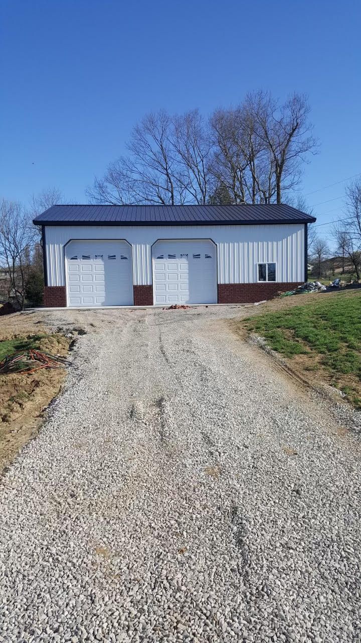 A white metal garage with two white doors and a brick base, sitting at the end of a long gravel driveway under a blue sky.