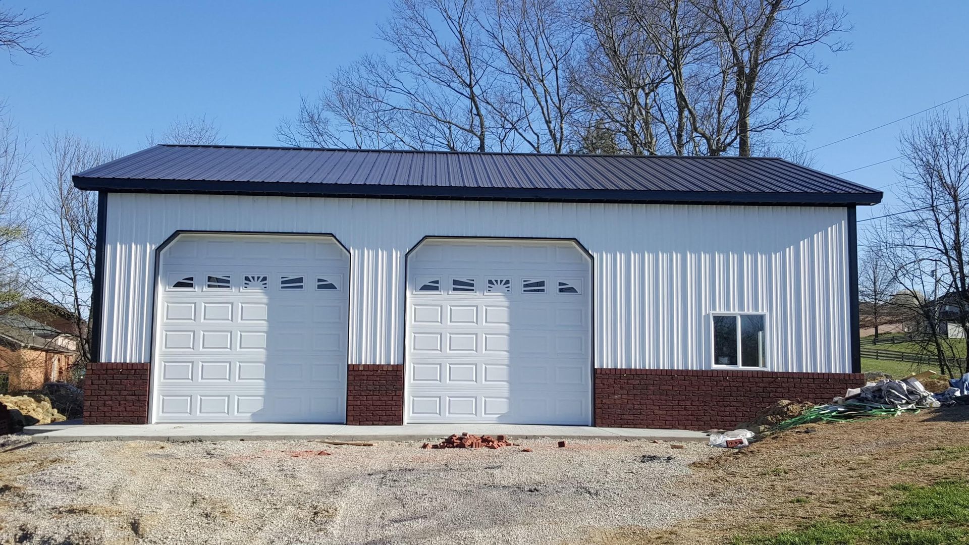A white metal two-car garage with a dark roof, brick-patterned base, and a side window, set in a sunny outdoor area.