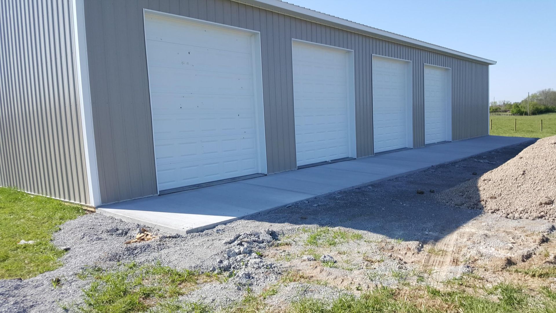 A large, grey metal building with four white garage doors, situated on a concrete slab next to a gravel path and grass.