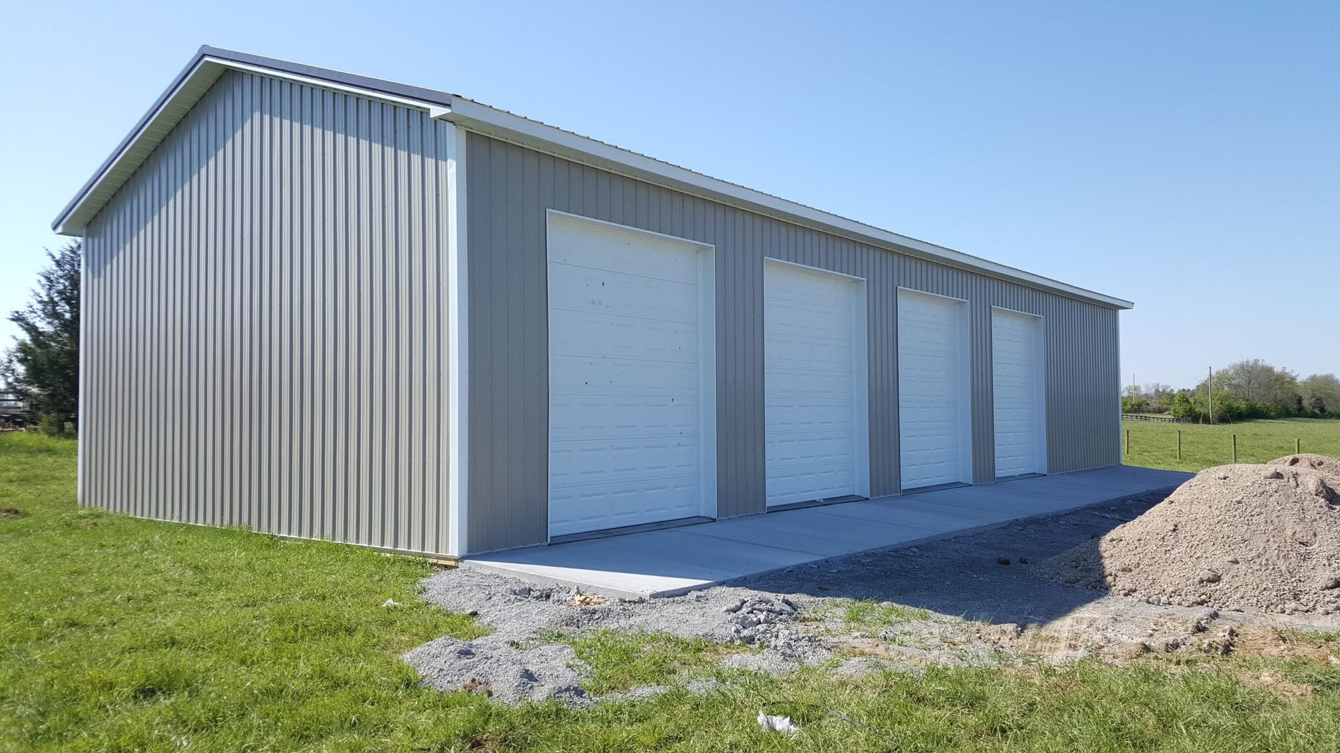 A light gray metal garage with four white roll-up doors stands in a grassy field under a clear blue sky.