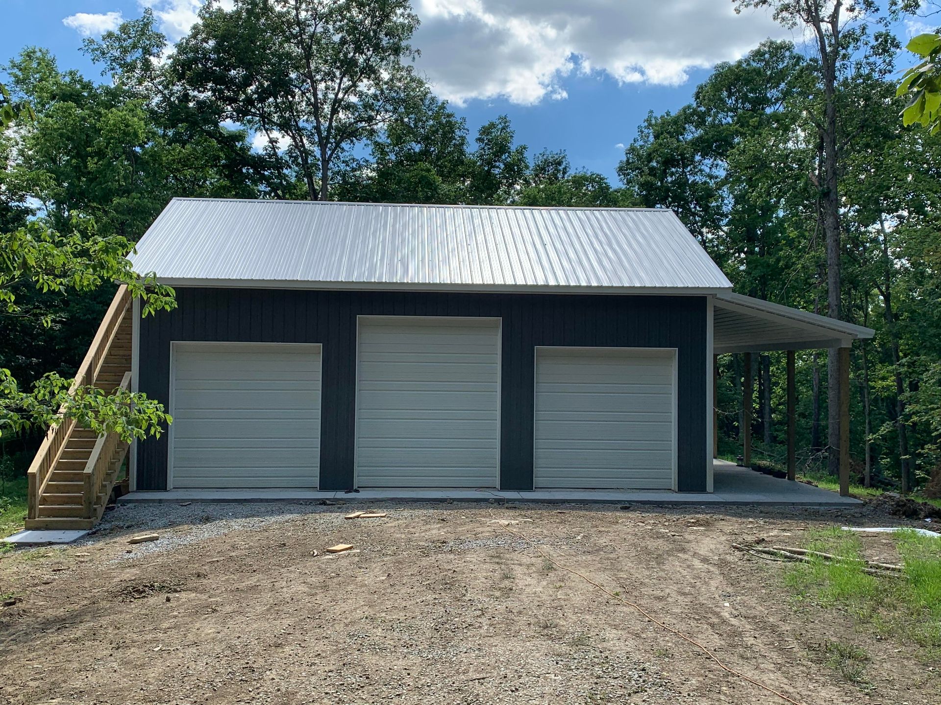 A dark grey garage with three garage doors, a silver metal roof, and an exterior wooden staircase, set in a wooded area.