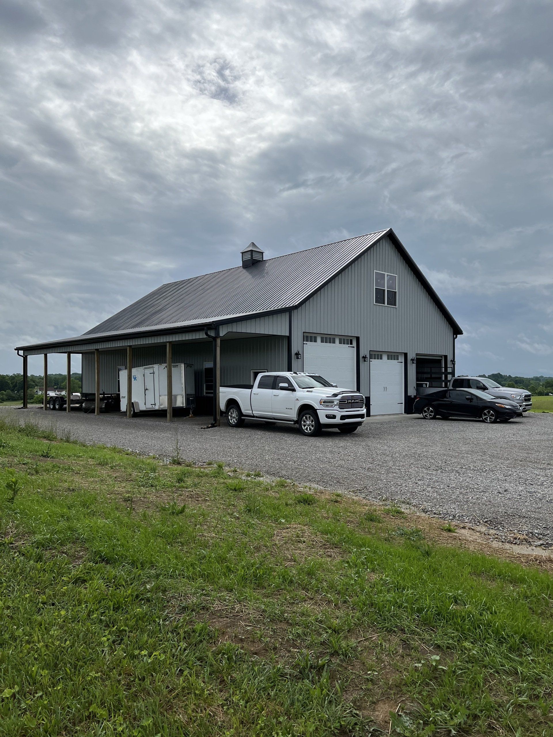 A light gray pole barn with a unique swirling pattern on the roof, surrounded by a gravel lot, grass, and vehicles.