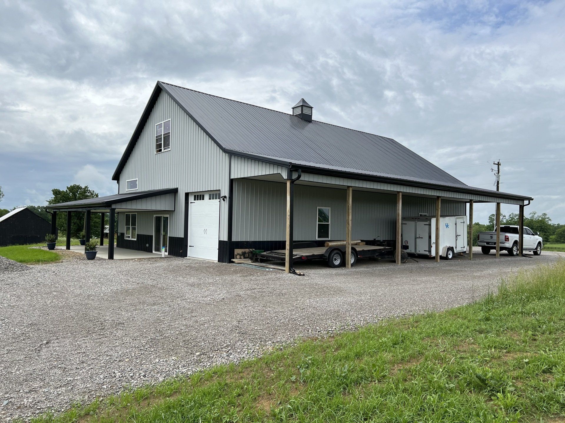 A light-gray pole barn with a dark metal roof and a covered porch, set on a gravel driveway under a cloudy sky.