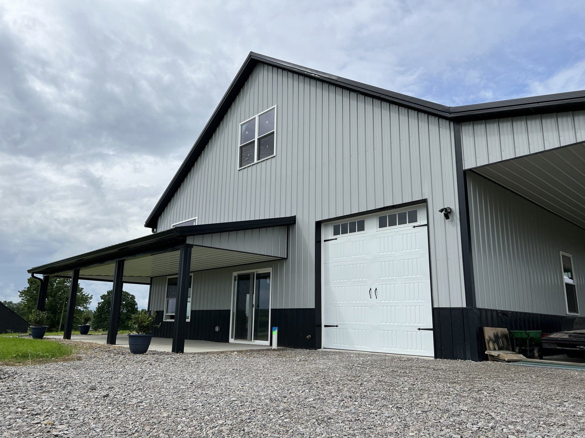 A grey metal pole barn with a black lower trim, a white garage door, and a covered porch on a gravel lot under a cloudy sky.