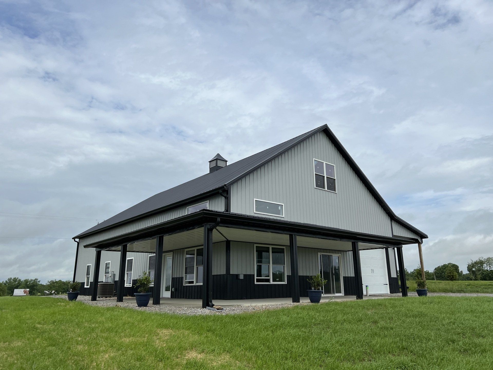 A modern gray and black barn-style building with a wrap-around porch set in a grassy field under a cloudy sky.