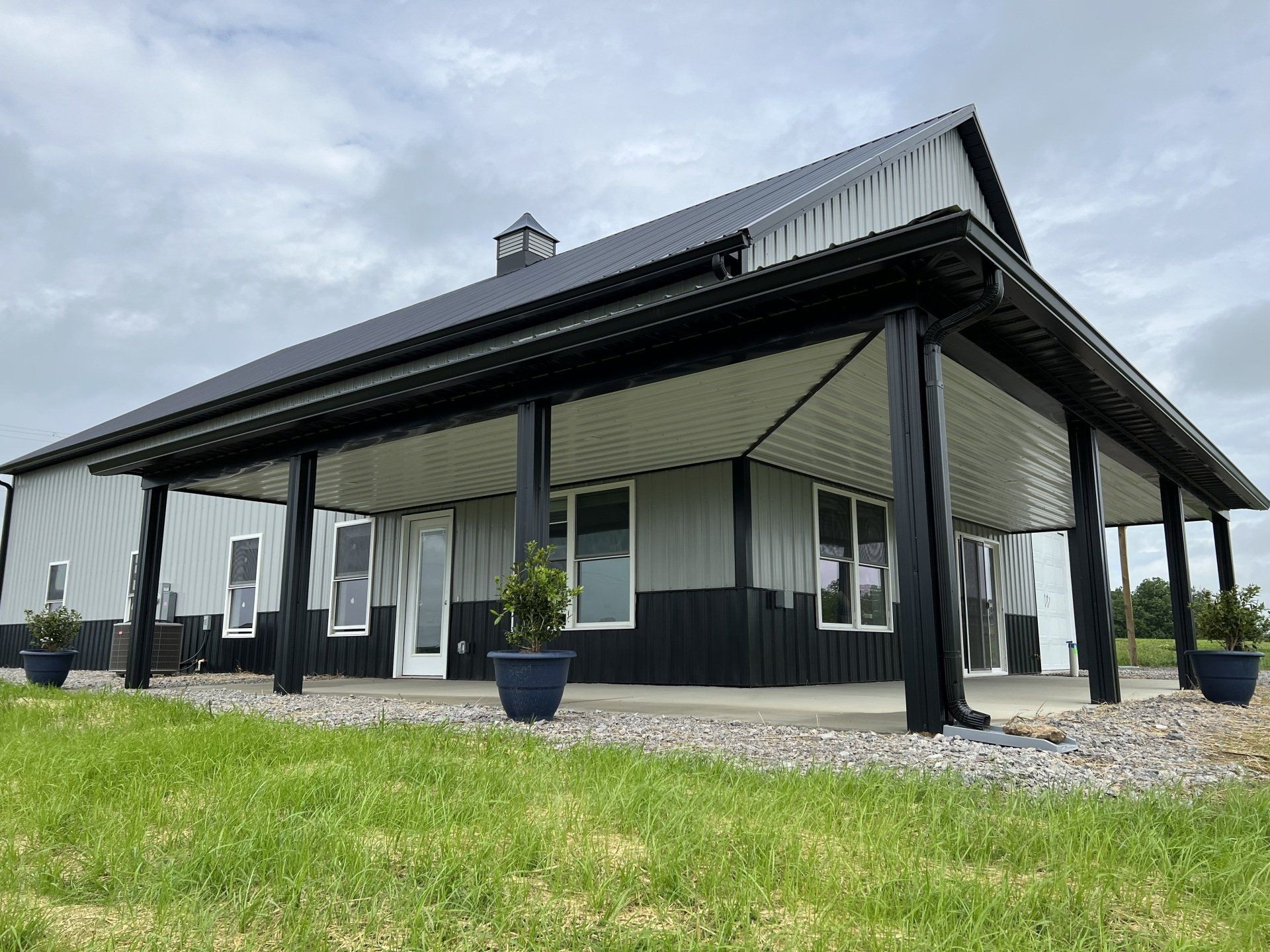 A modern farmhouse with a grey and black metal exterior, a large covered porch, and potted plants on a gravel ground.